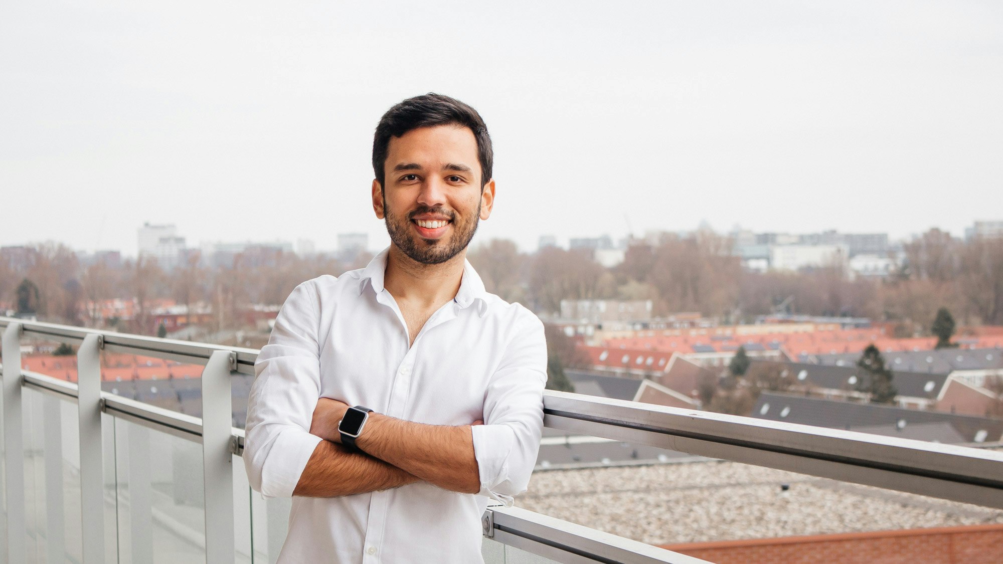 Man with dark hair, in white shirt on a balcony with a view over a city.