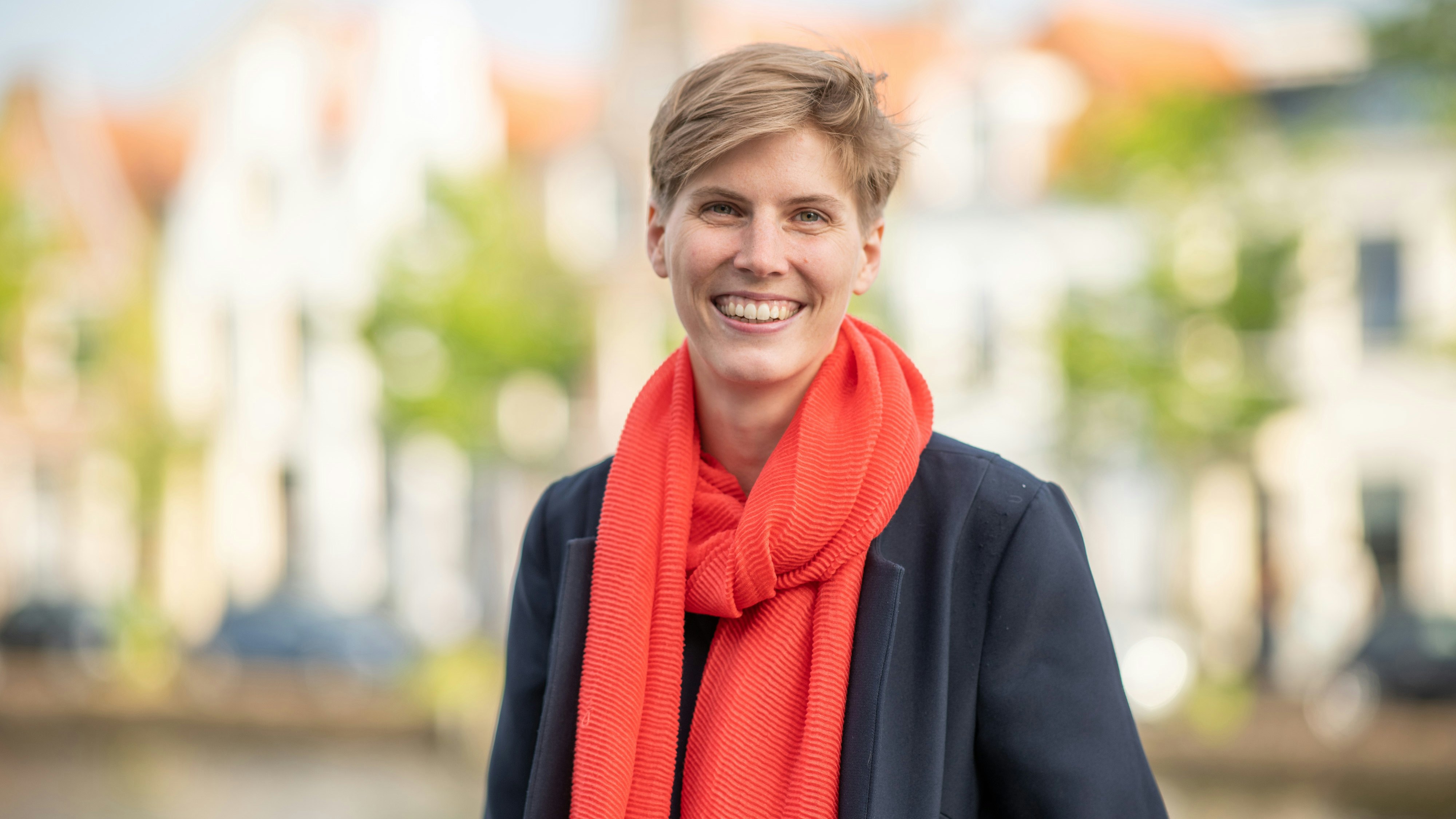 A smiling person with short hair, dressed with a dark coat and a red scarf.