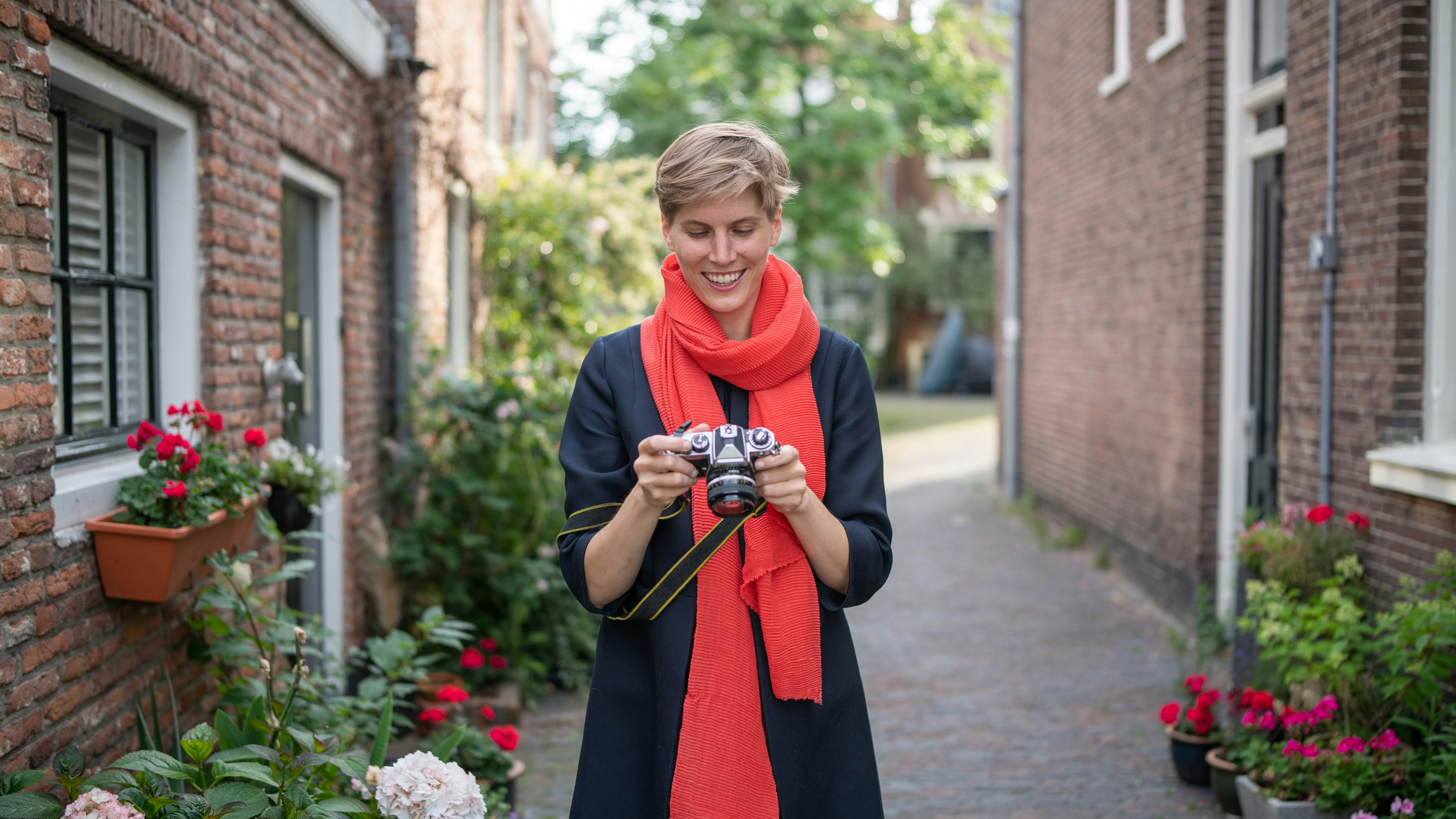 Annegret Bönemann, die einen leuchtend roten Schal trägt, blickt auf eine analoge Kamera, die in den Straßen einer alten Stadt steht.