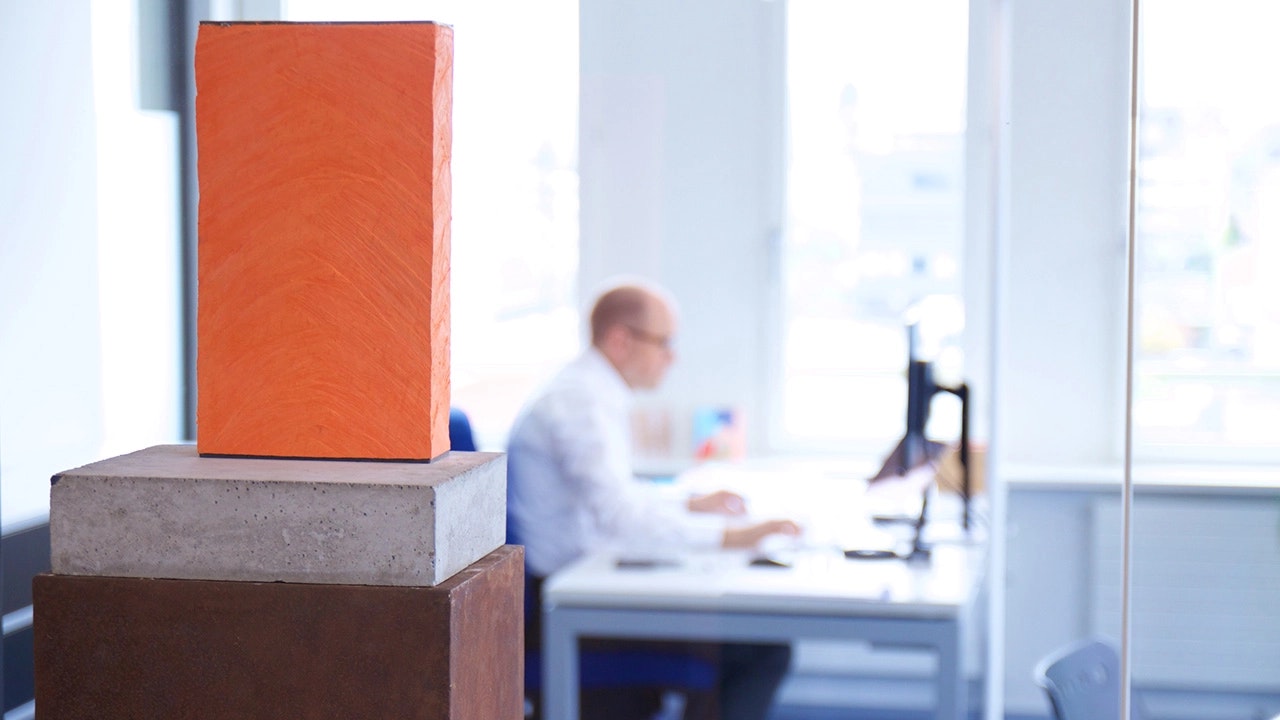 In the foreground an orange sculpture. In the background a person at work.