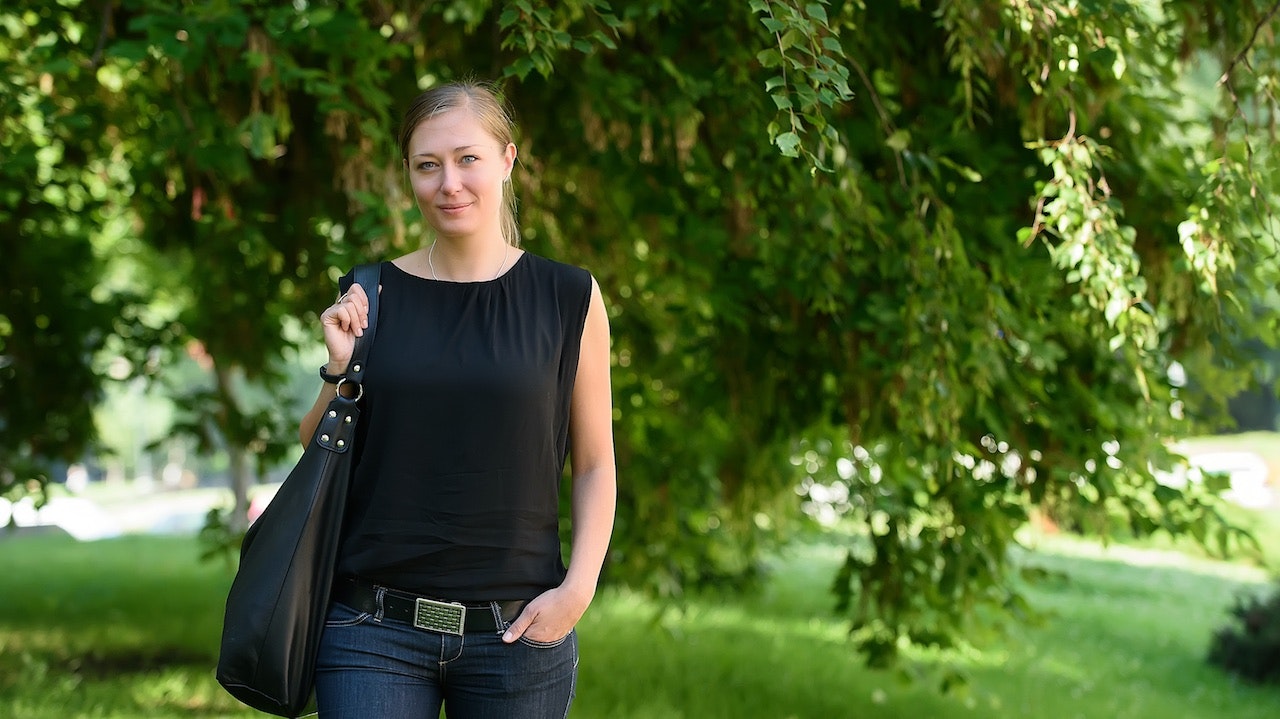 Albena is standing outside, holding her black handbag under a tree.
