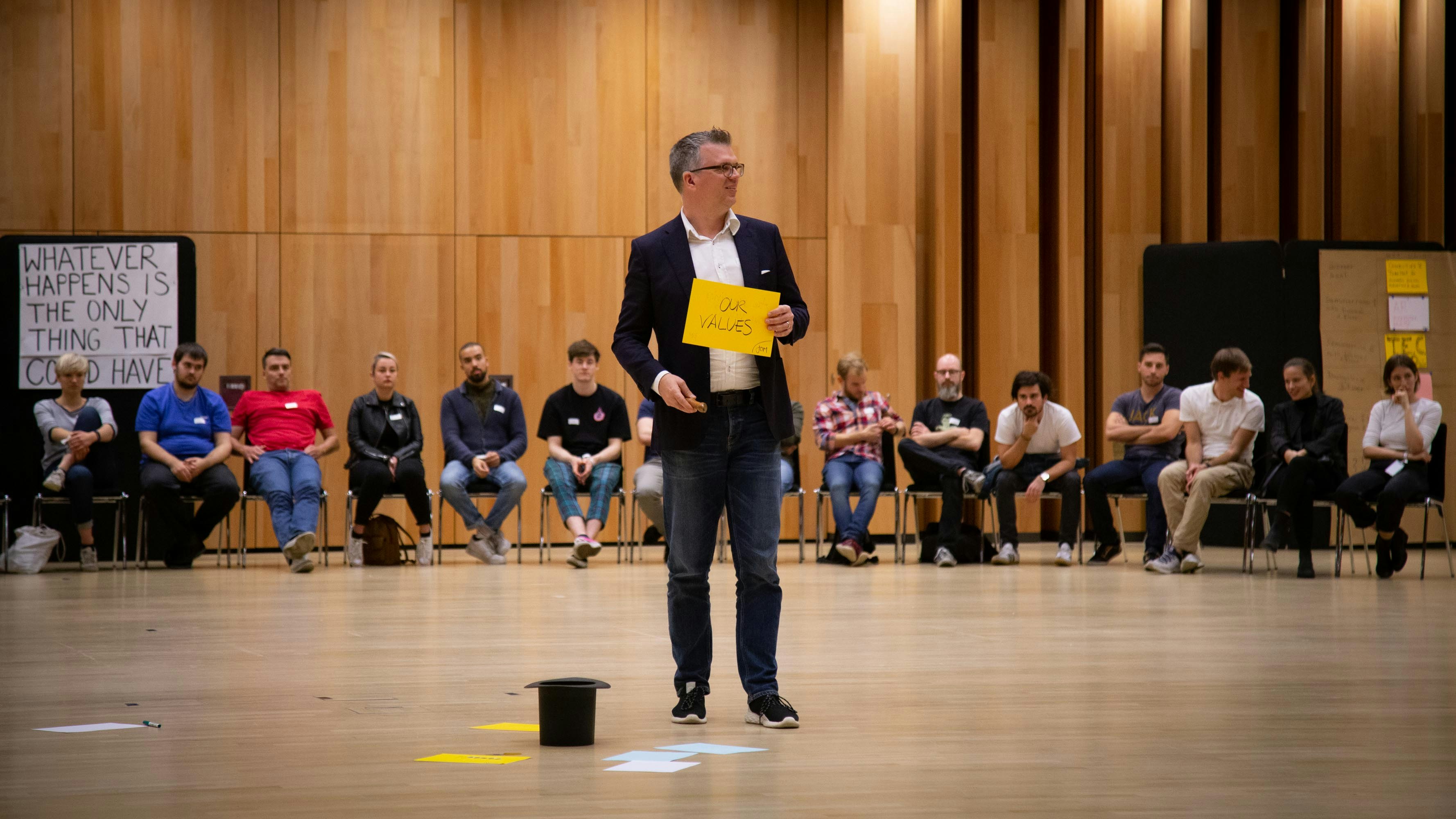 Jonathan Möller steht vor sitzenden Menschen und hält ein gelbes Papier "Unsere Werte" hoch.