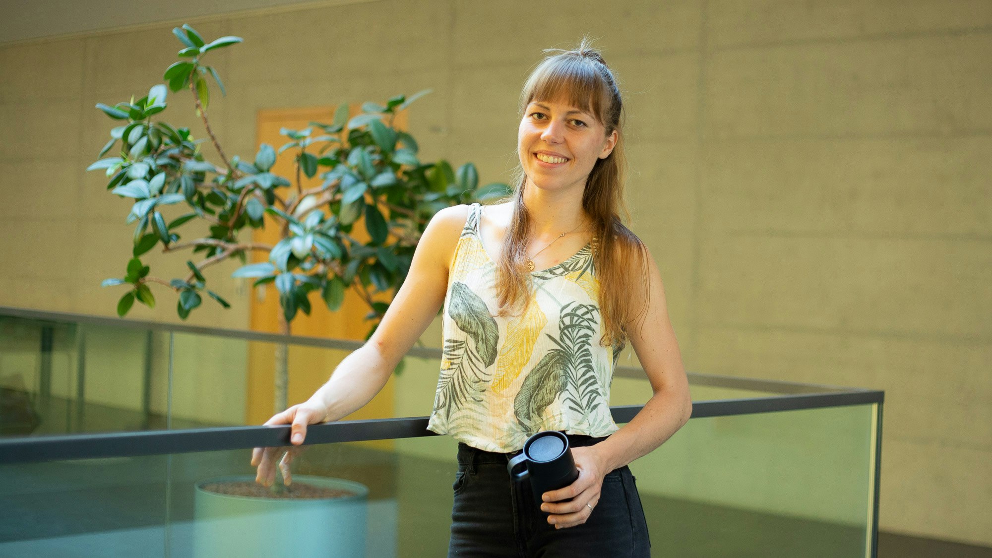 Johanna Eppler, with long blonde hair, wearing a shirt with a plant motif, grips a banister in a stairwell. In the background a plant.