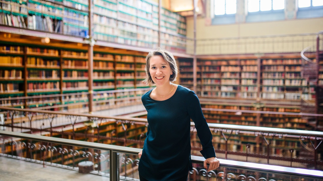 Valerie is standing in a library, full of books in wooden shelves n a balcony.