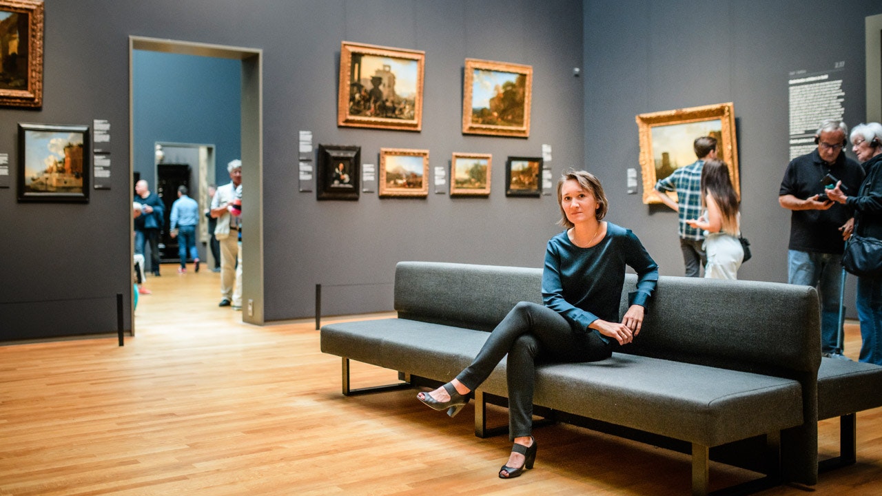 Valerie is sitting on a grey couch in an art gallery.