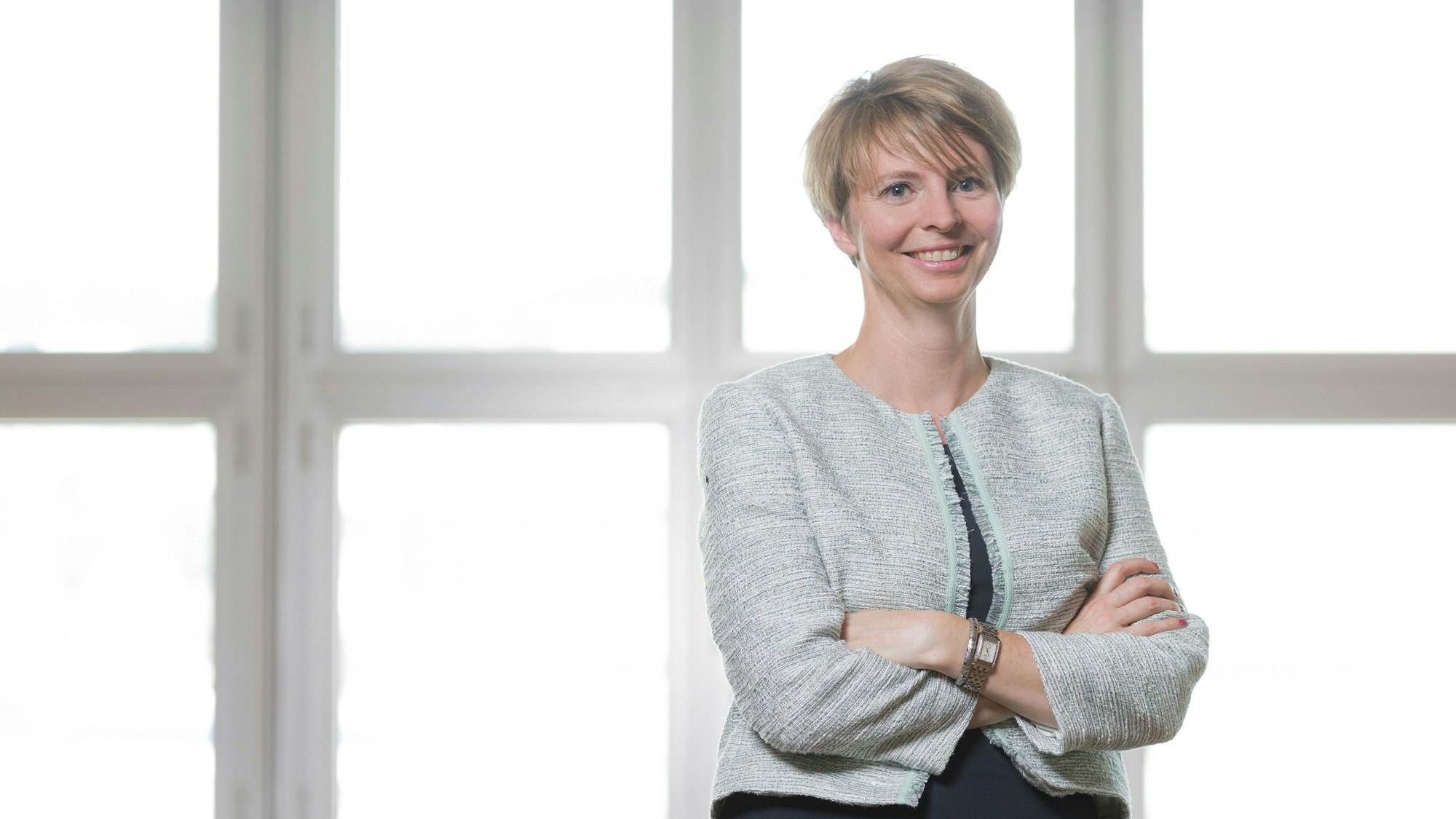 Jeannine steht mit verschränkten Armen, weißes Fenster im Hintergrund.