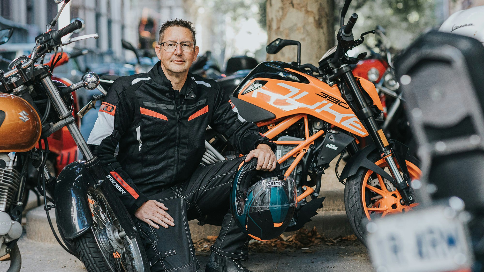Mick in a dark outfit, in front of an orange motorbike, helmet in hand.