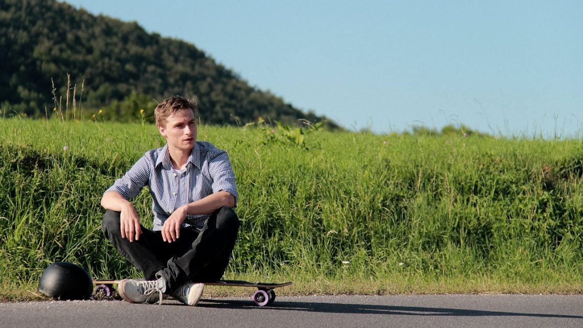 Claus Stachl sitting on his skateboard on a road through nature.