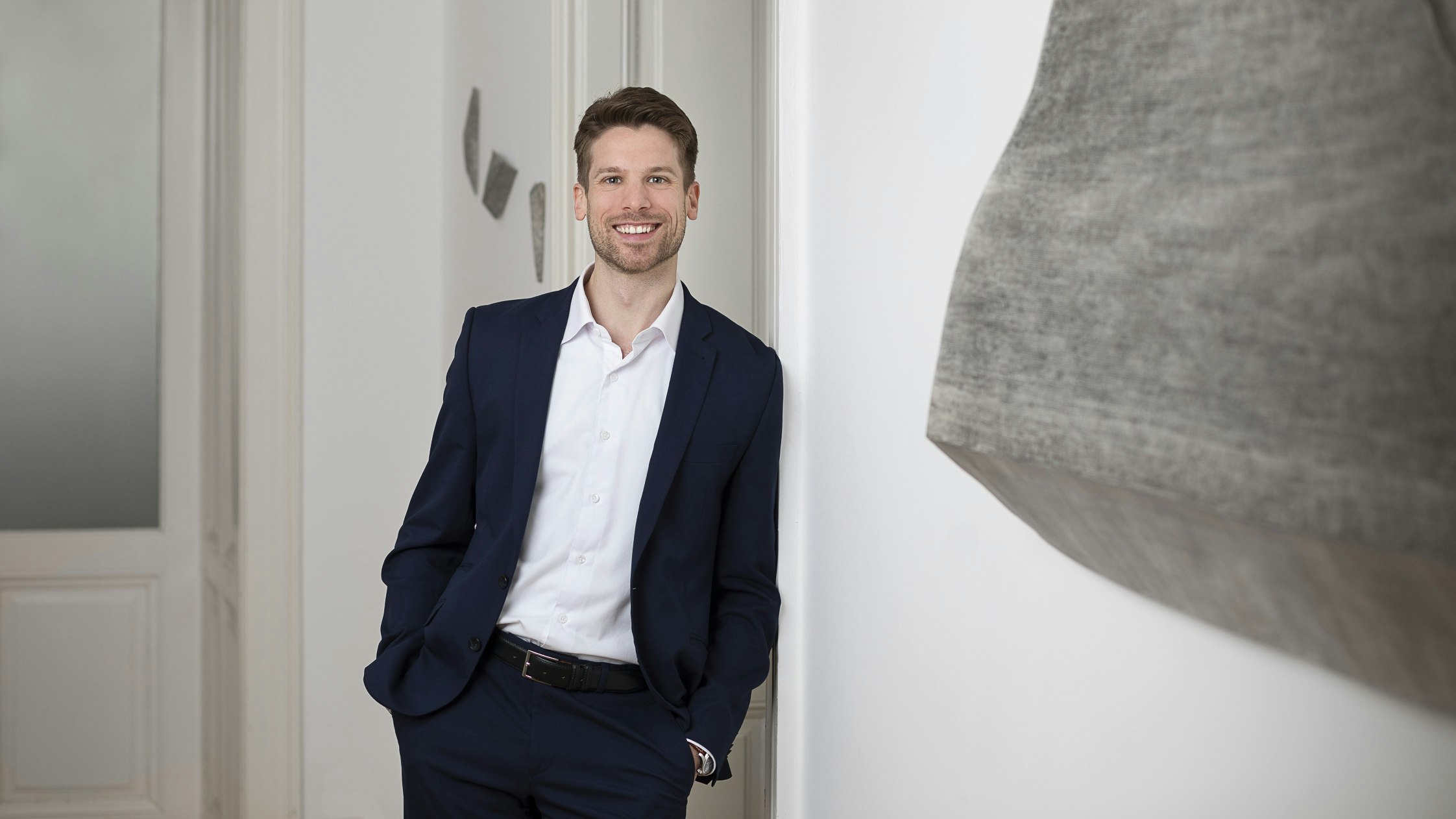 A man in a suit, smiling in an office with art on the wall.