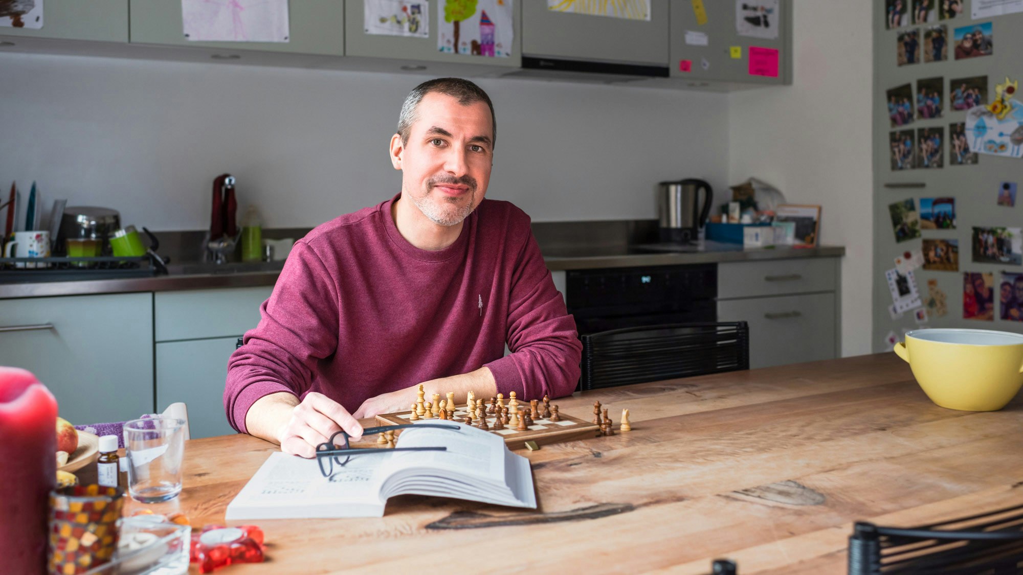 Christian Wymann sitzt an einem Küchentisch mit einem Schachbrett und einem Buch vor sich.