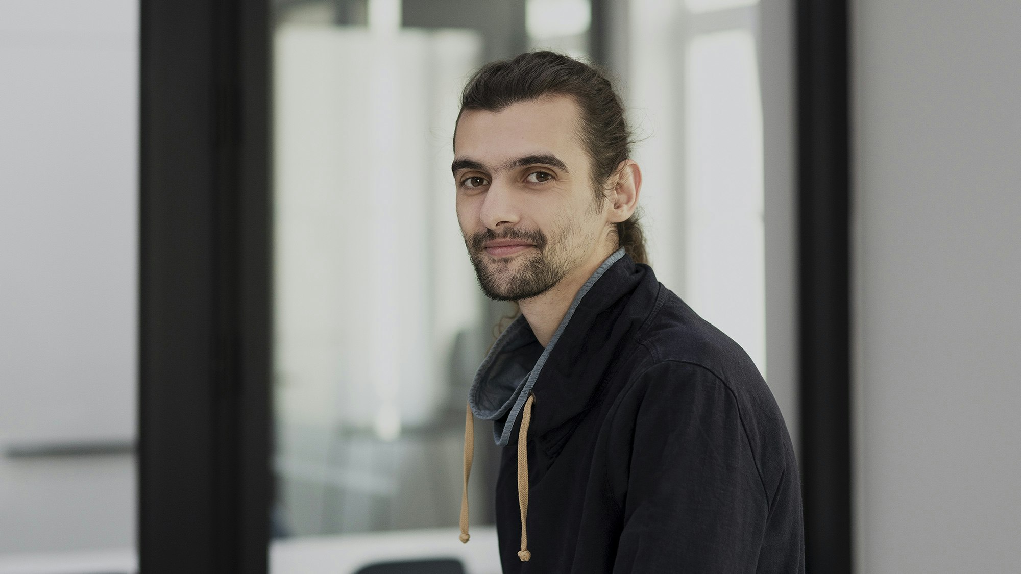 Man mit langem dunklen Haar in schwarzem Pullover lächelt in die Kamera.