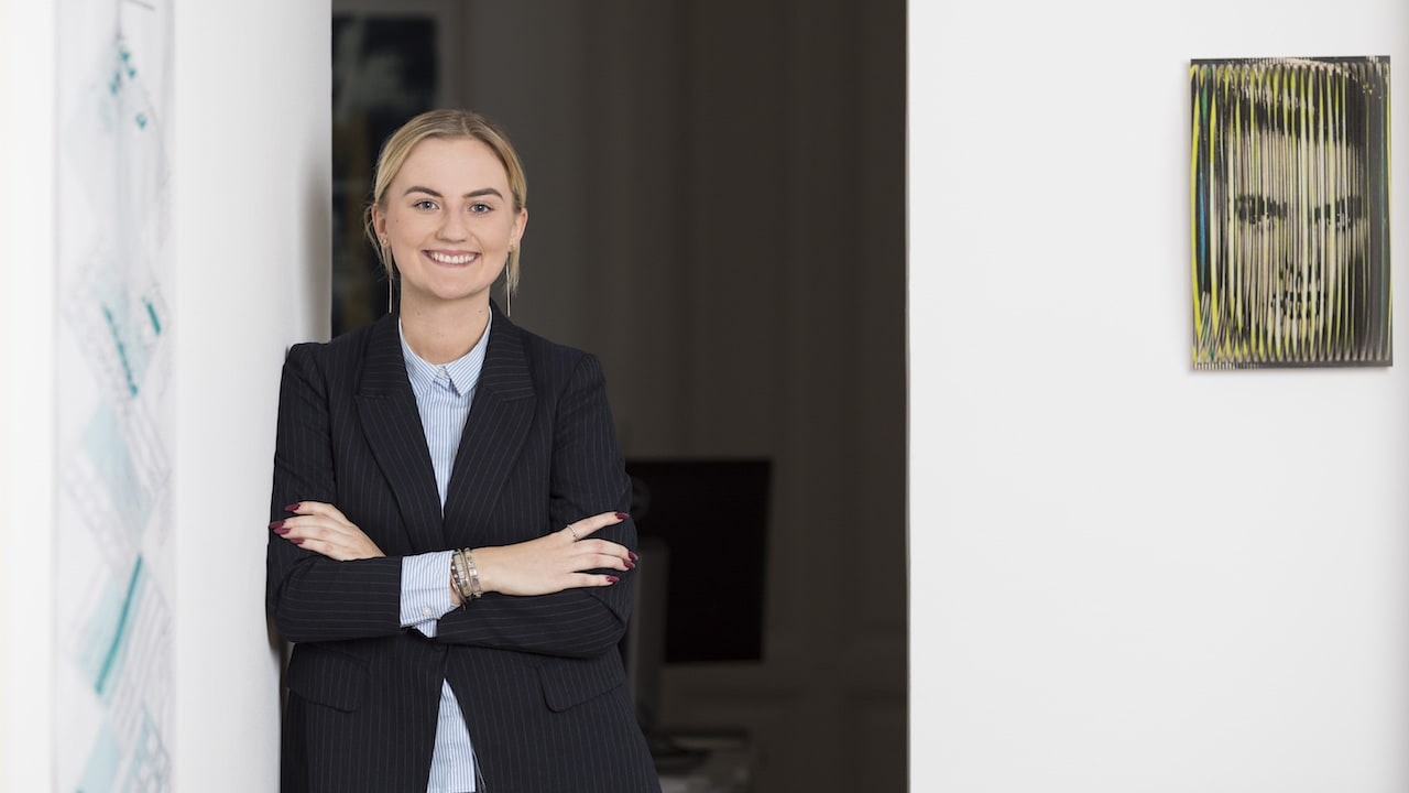 Jacqueline mit blondem Haar, in schwarzer Jacke, lehnt an einer Wand. Rechts ein Gemälde.