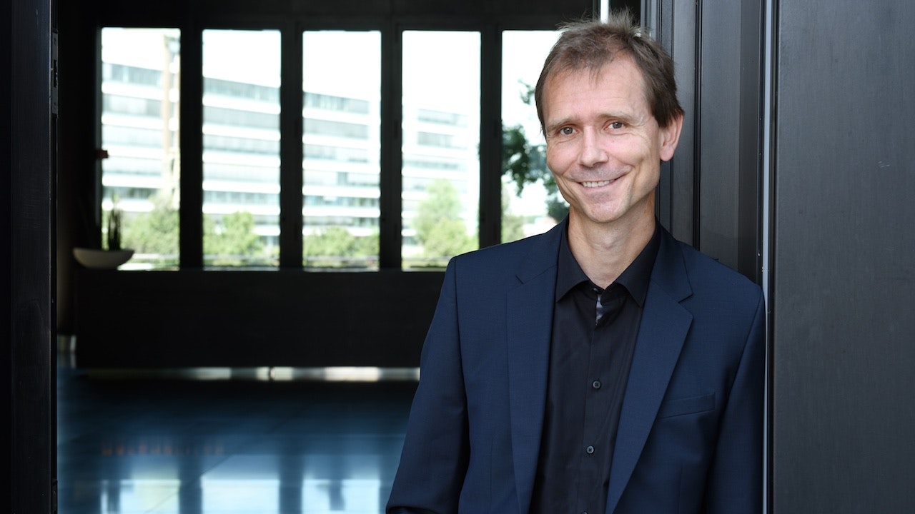 Man in dark blue suit leans against a wall and smiles at the camera