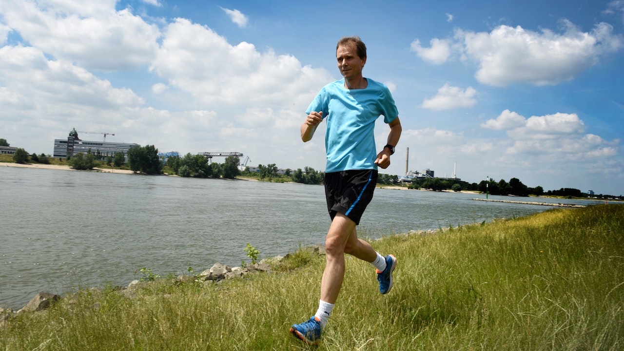 Andreas Drexhage in einem blauen Hemd, der im Gras an einem Fluss entlang läuft.