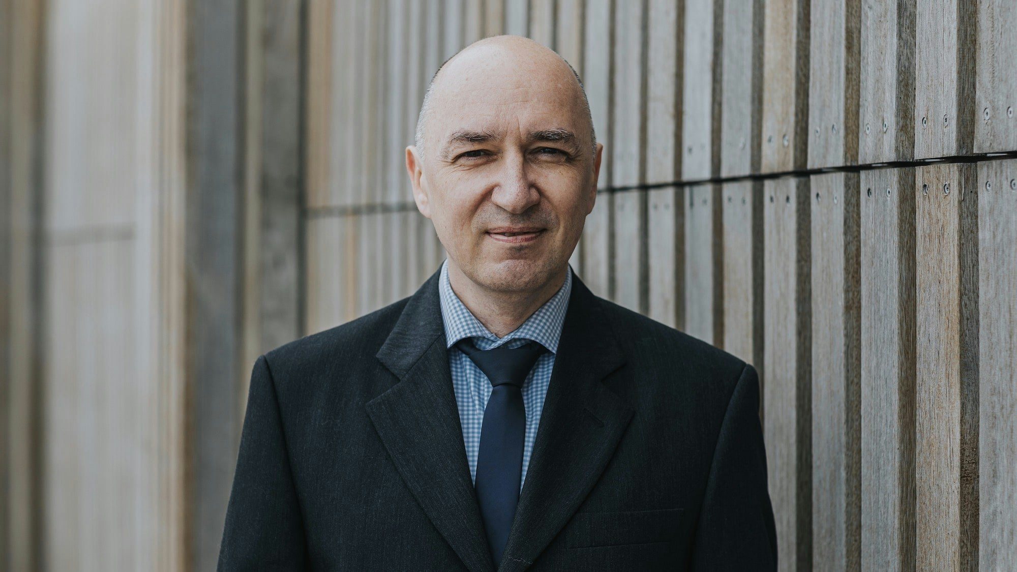A man without hair in a dark suit in front of a wooden wall.