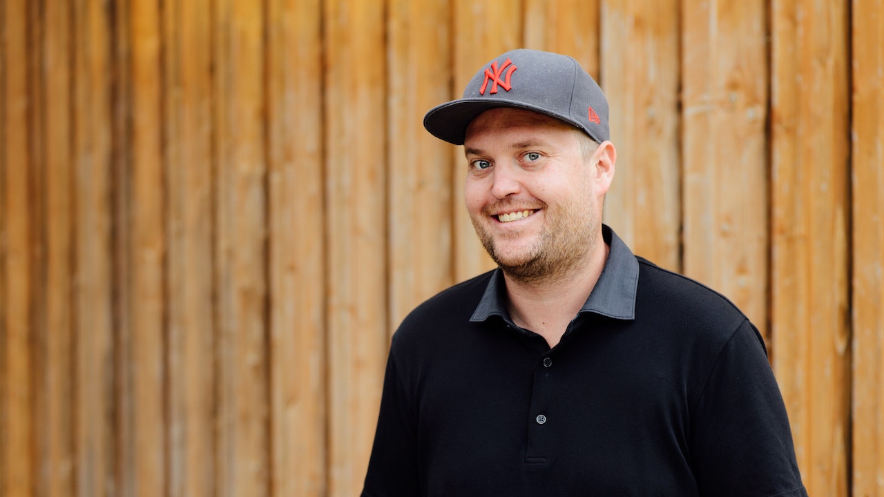 Florian with a cap in a black polo shirt, in front of a wooden wall, smiling into the camera.