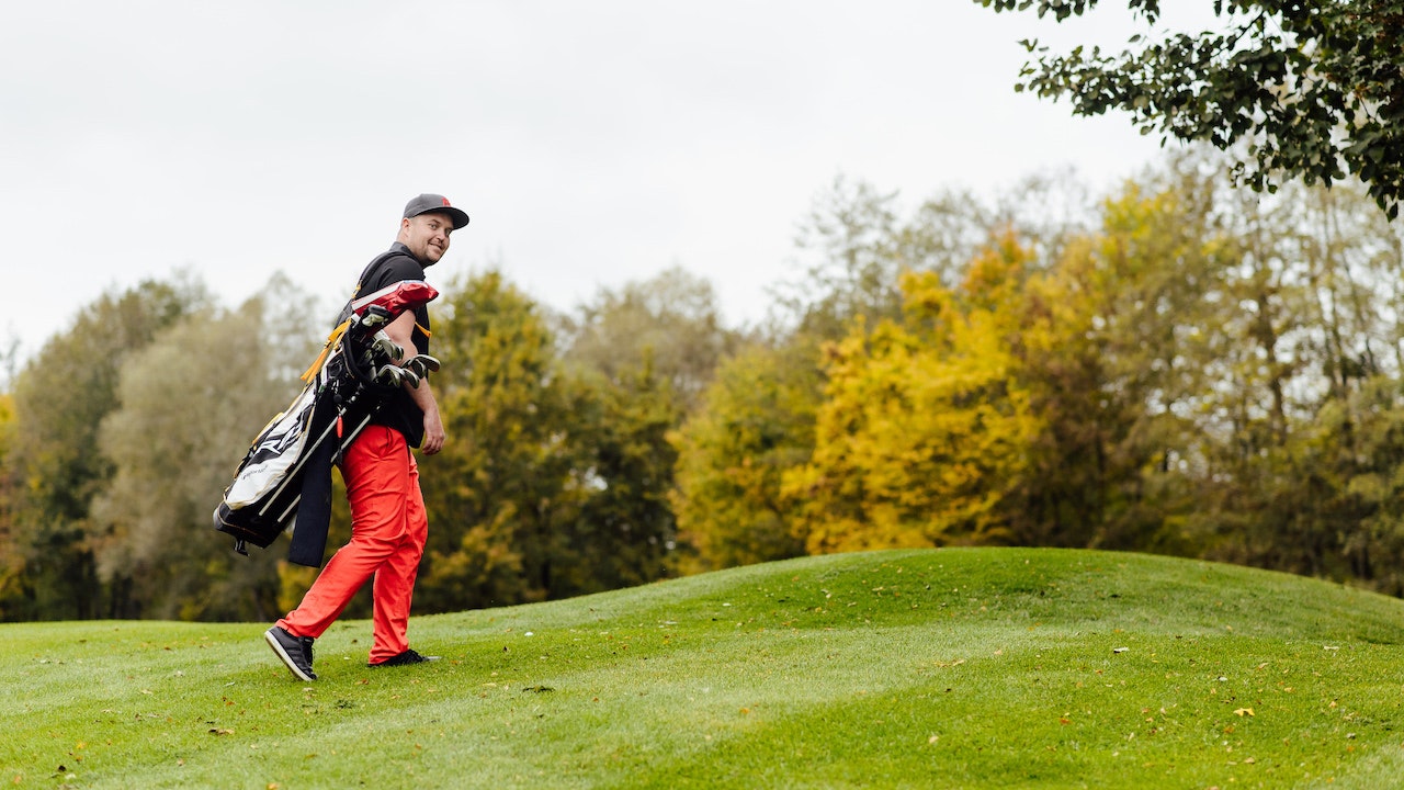 Florian in red trousers, with cap, carrying golf equipment on a green lawn.