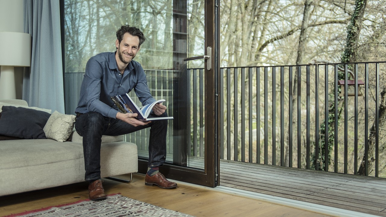 Sebastian sitting on a couch, looking in the camera, holding a book open. In the background a balcony in the green.