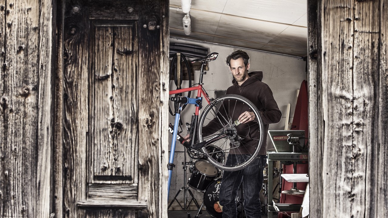 Ein Mann schaut in die Kamera, während er in einer Garage ein Rennrad hochhebt.