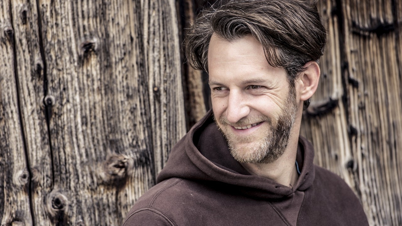 Man smiling, in brown jumper in front of wood.