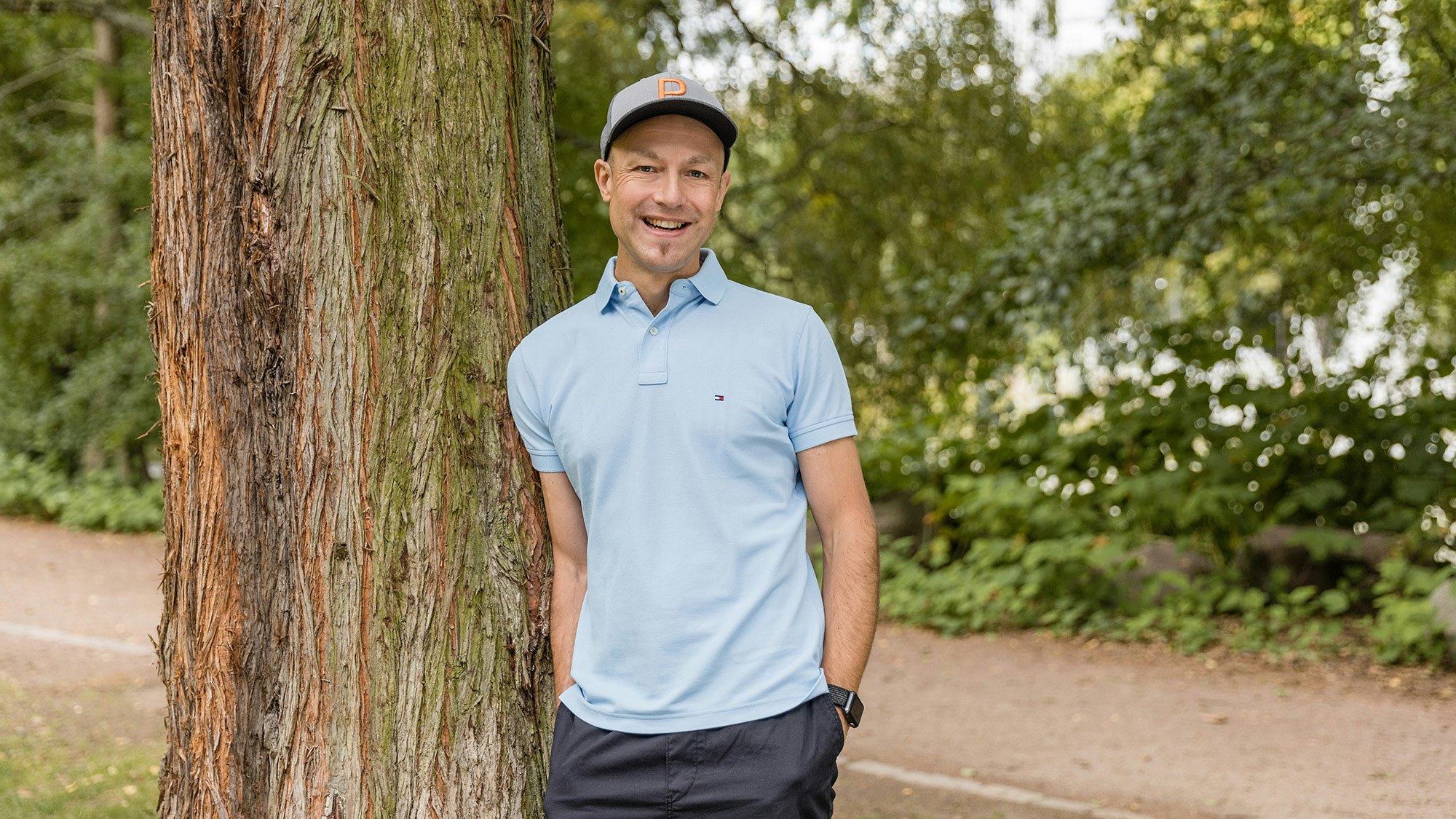 Patrick leaning against a tree, wearing a blue shirt and cap, smiling