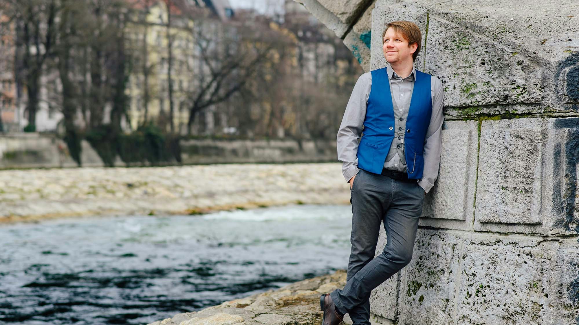 Dennis van der Wals is standing on a river bank, leaning against a stone bridge pillar with his hands in the pockets.