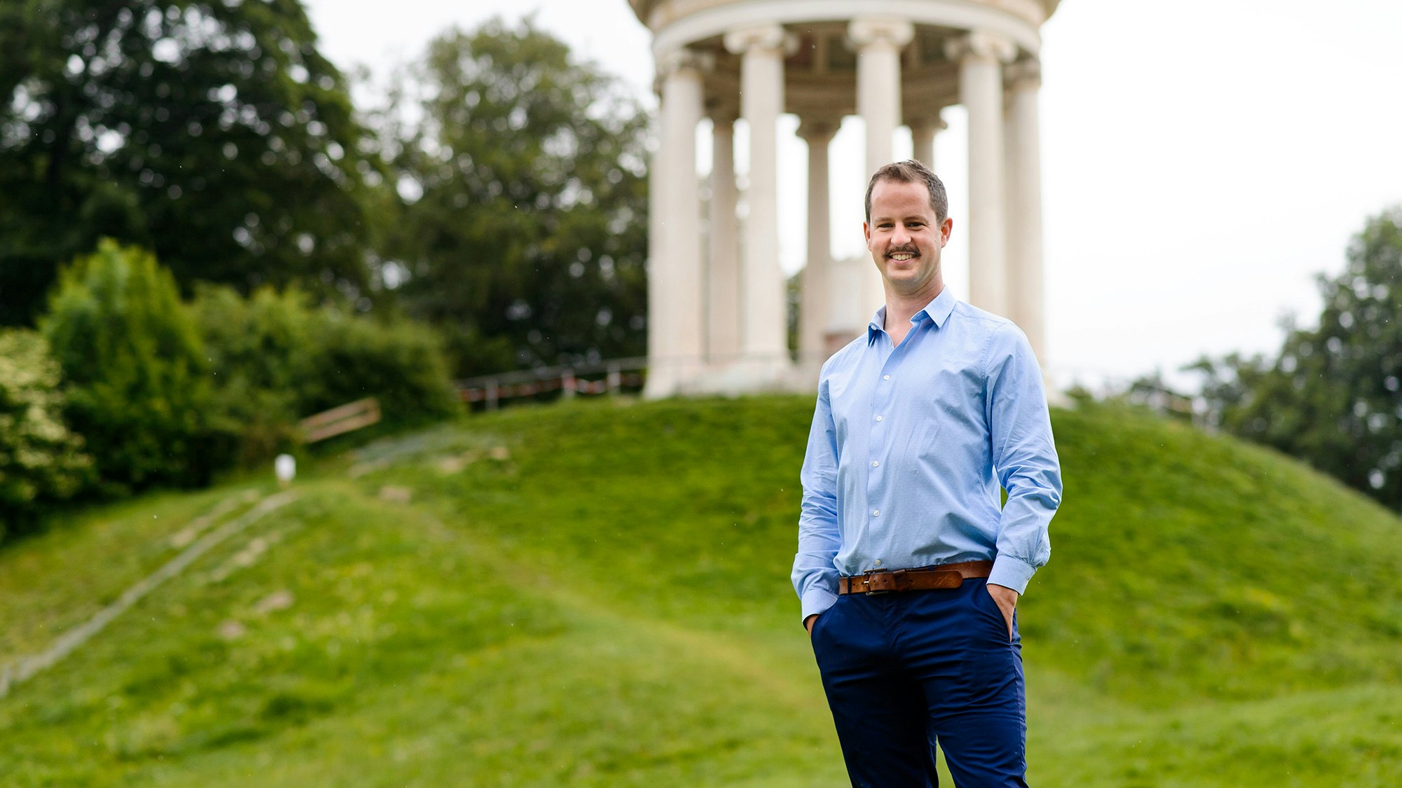 Carl Simbruner in a blue shirt and formal trousers is standing in a park with a white stone gazebo behind him.