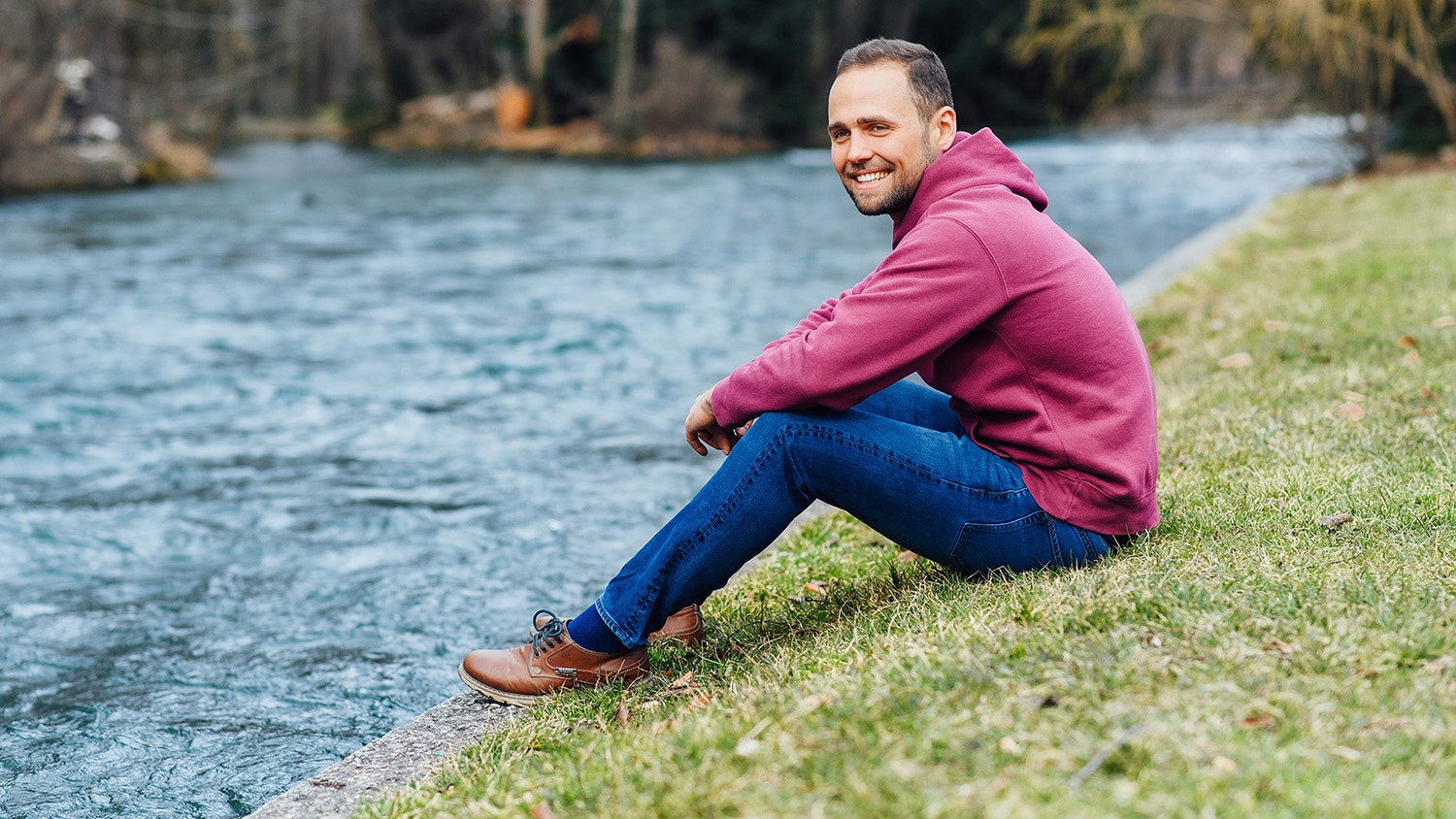 Tobias Bartsch sitzt auf einer Wiese am Flussufer und lächelt.
