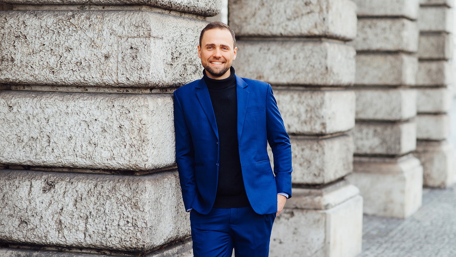 Tobias Bartsch is standing in marine blue suit with black sweater next to stone pillars.