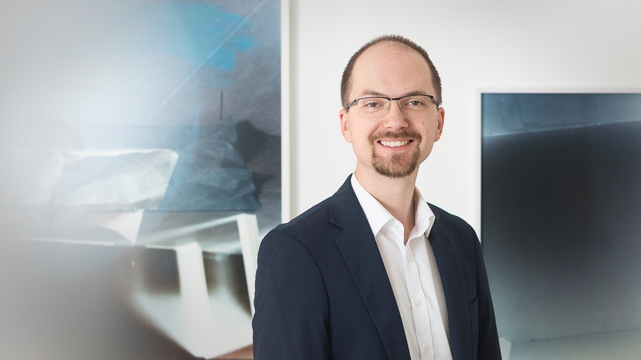 Profilfoto von Andreas Damberger im Anzug mit weißem Hemd mit dunkelblauer, verschwommener Kunst im Hintergrund.