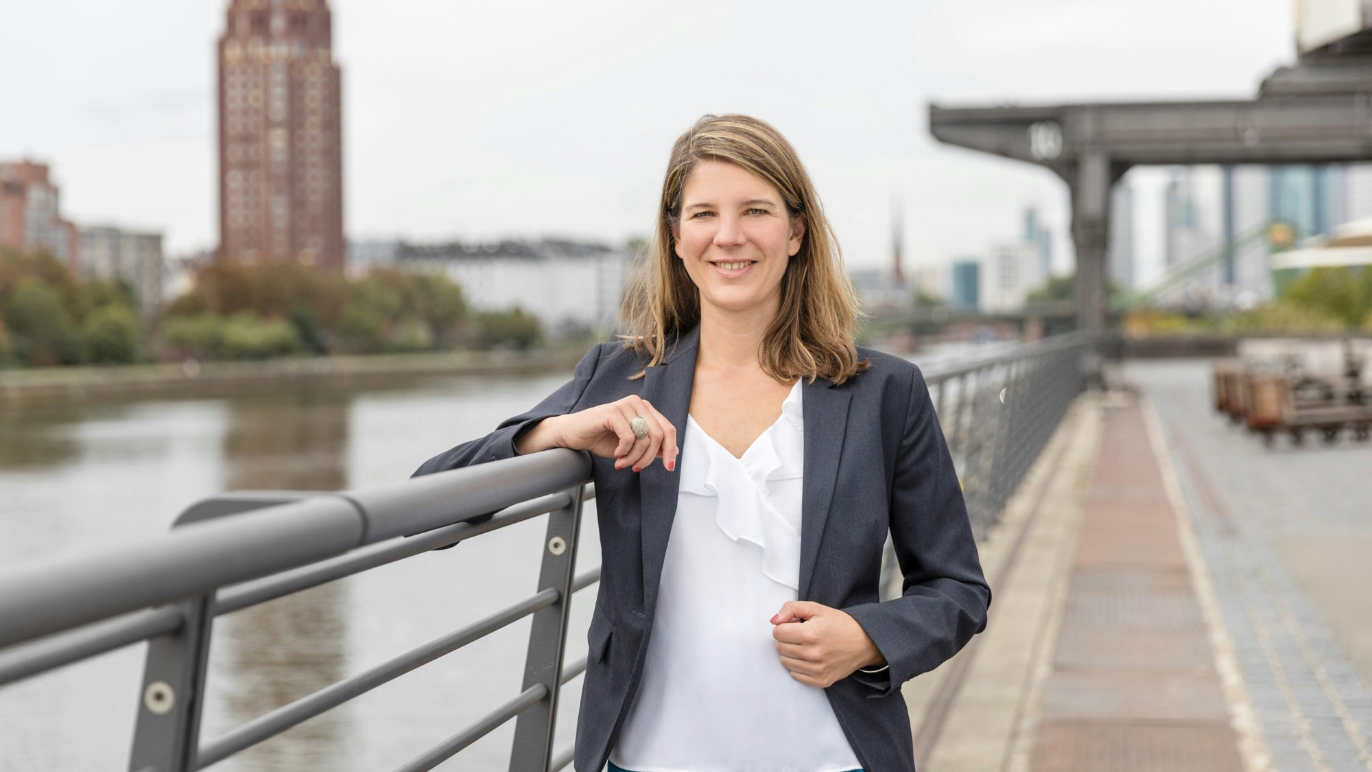 Isabel Schiller steht an einem hohen Geländer am Ufer eines großen Flusses in einer Stadt.