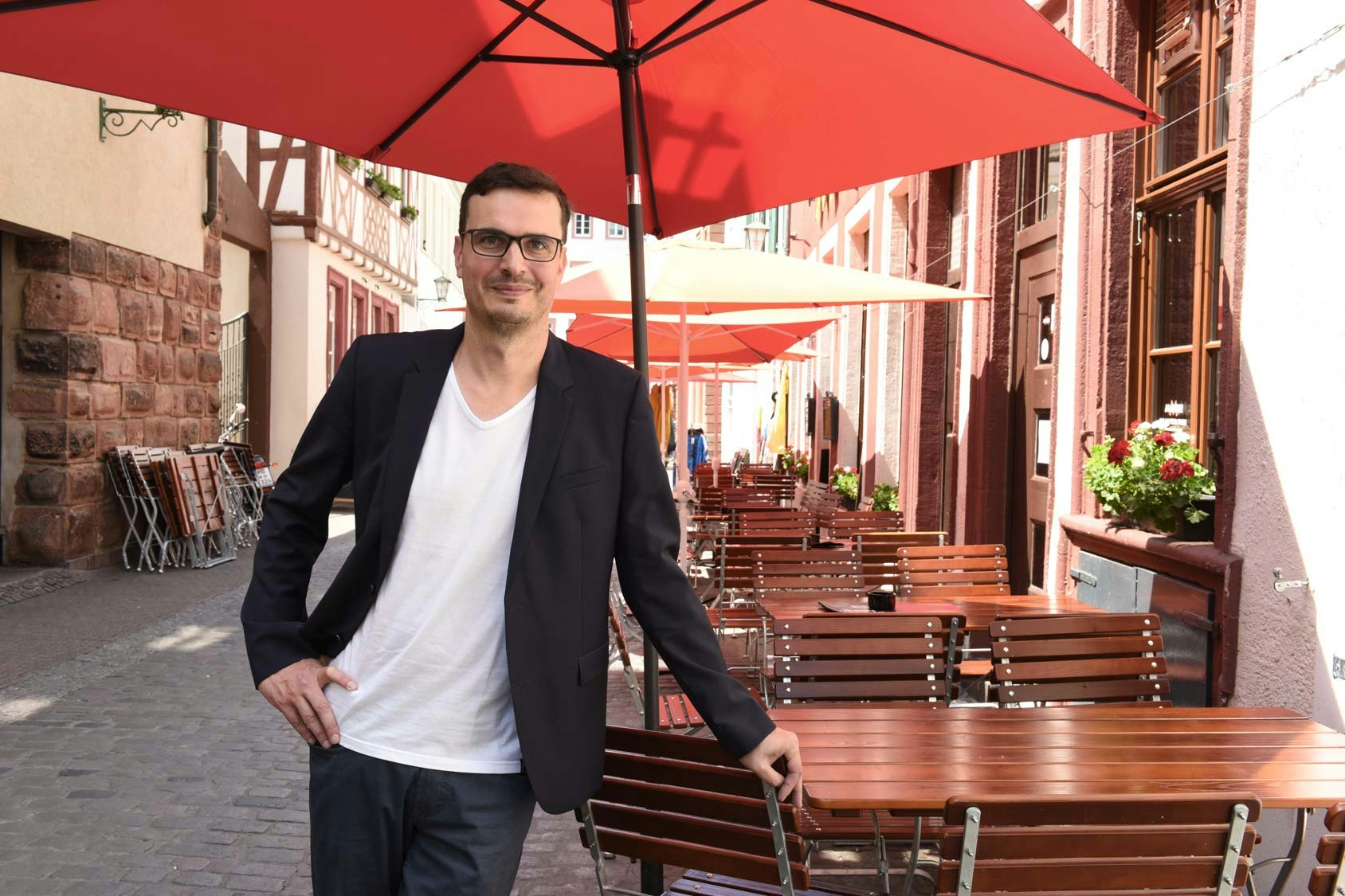 Christian Weih-Sum is leaning on the back of a wooden chair outside on a restaurant garden, under red umbrella.