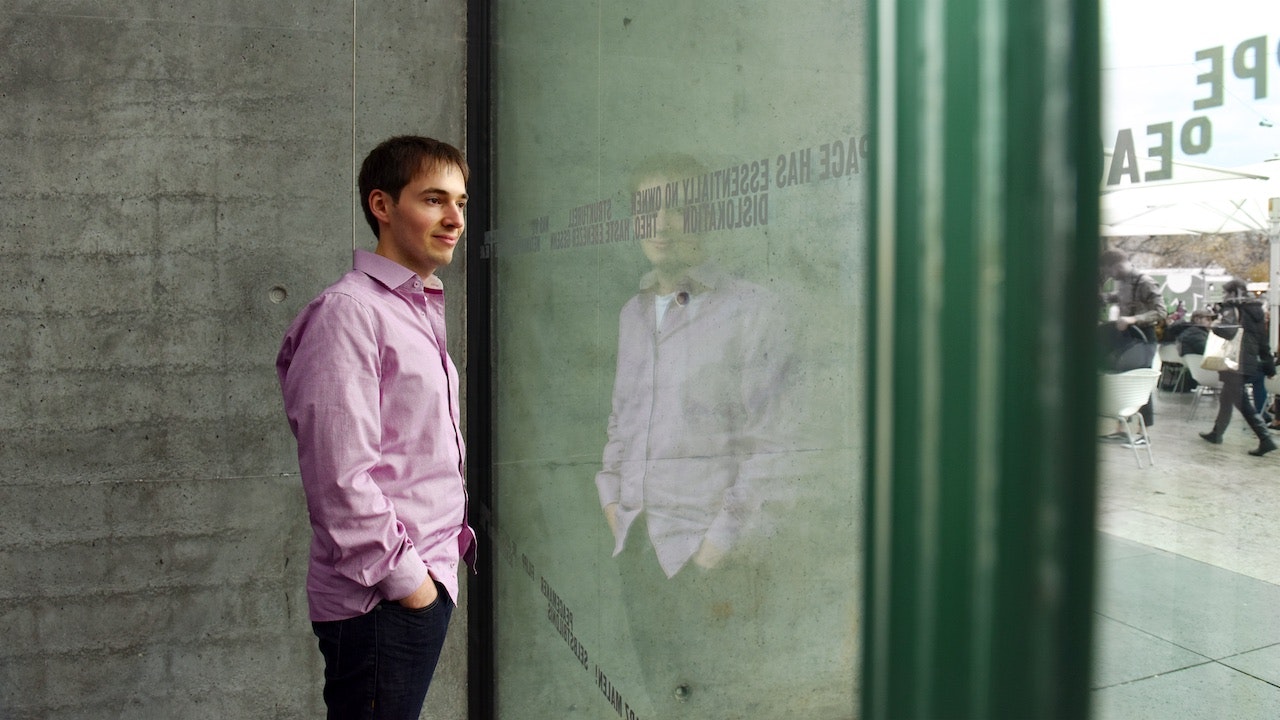 Daniel Kimmich, dressed in a pink shirt is standing with hands in his pockets, looking outside from a modern office.