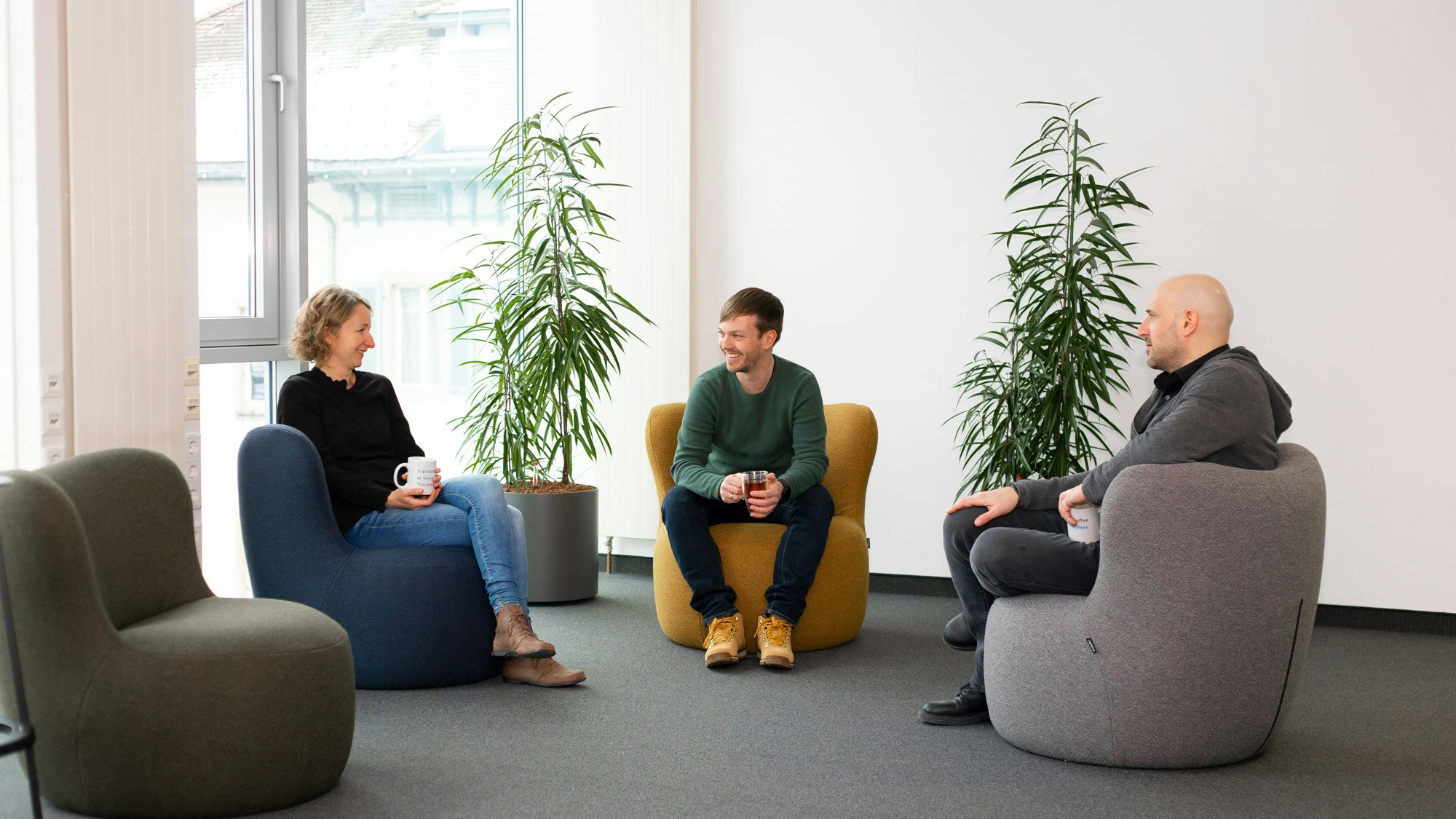 Three people sitting on comfy chairs in an office environment, surrounded by plants