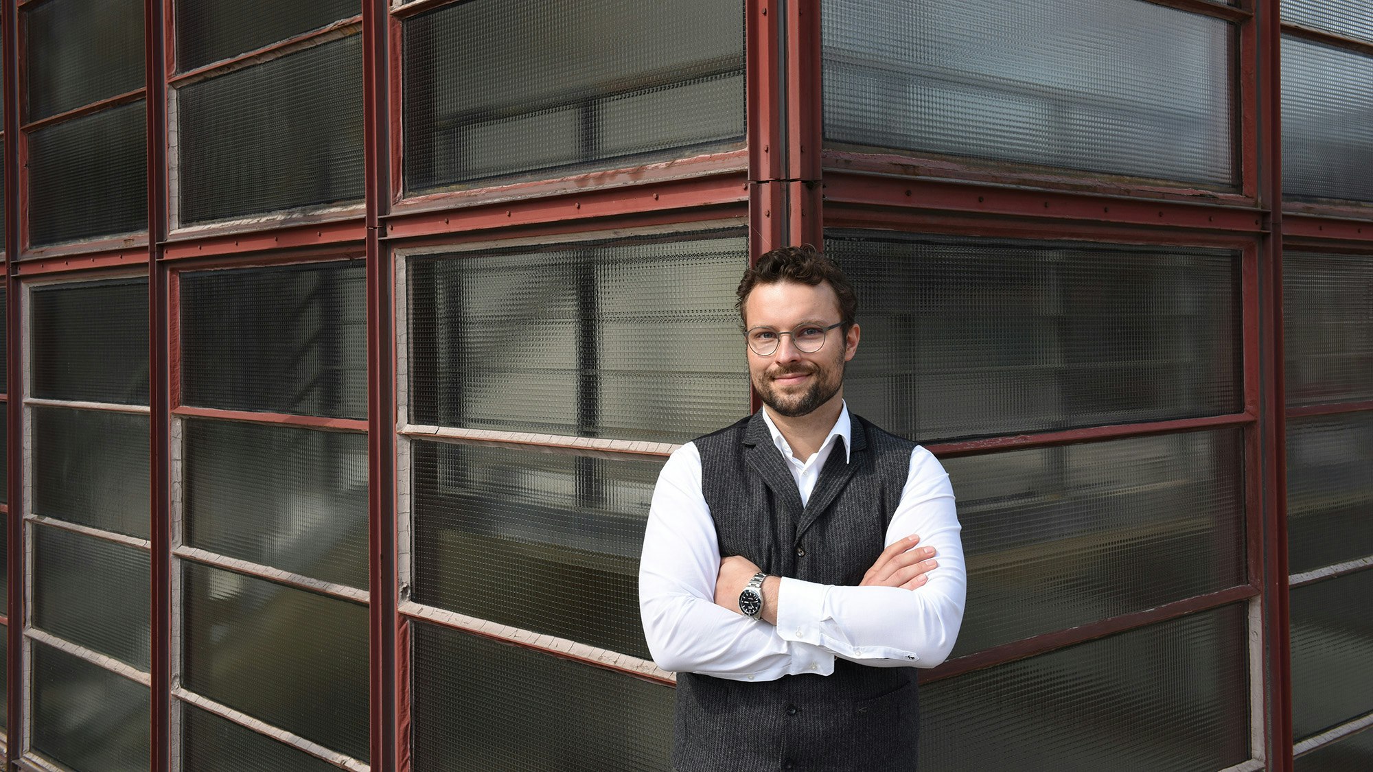 Person in glasses, standing at the corner of a building with arms folded.