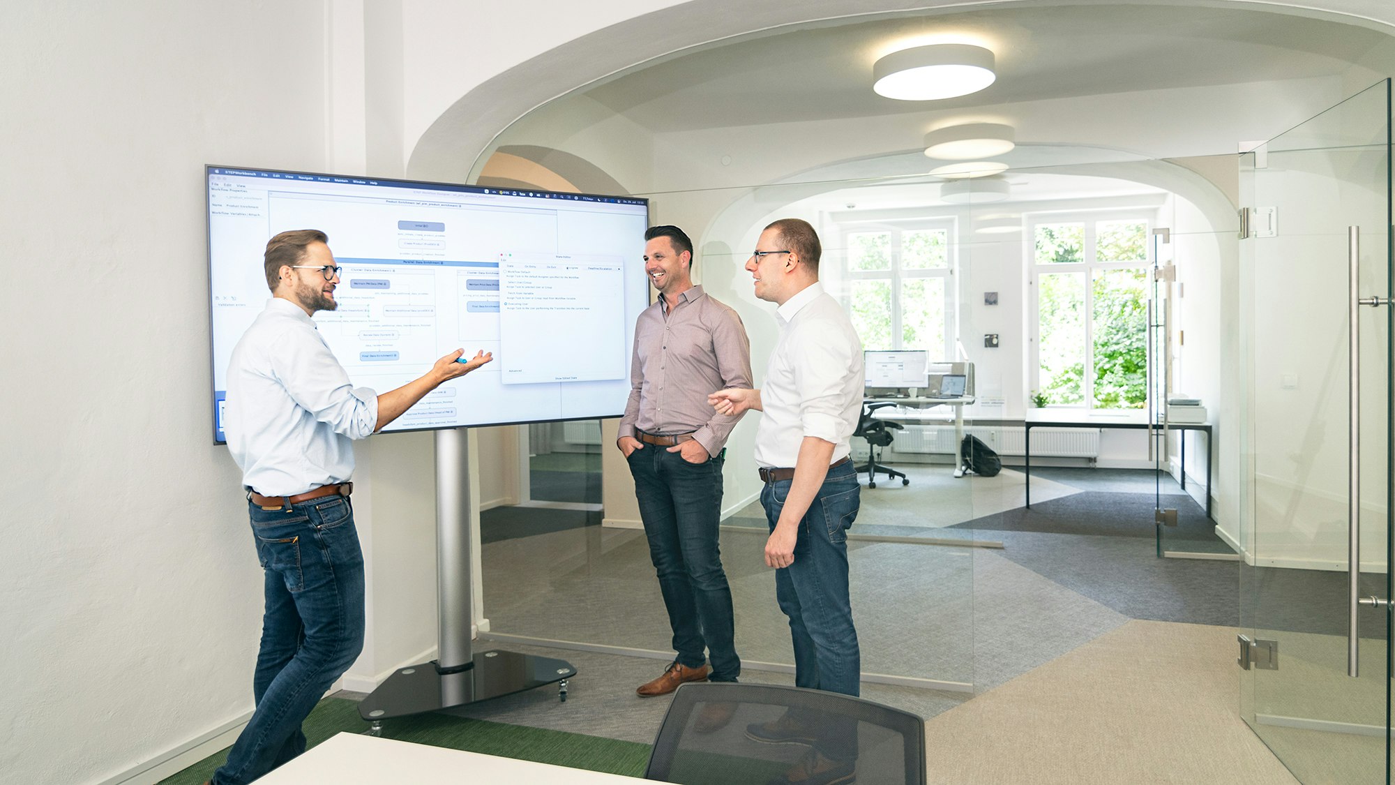 Drei Menschen diskutieren in einem Büro. Im Hintergrund ein großer Bildschirm.