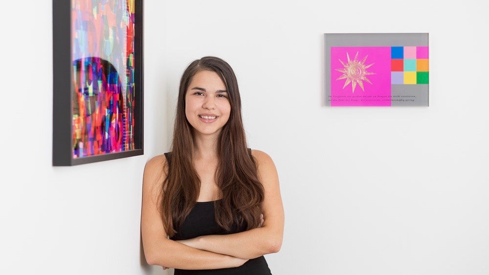 Sabine leans casually against the wall in the office. Two works of art in bright colors hang in the corner.