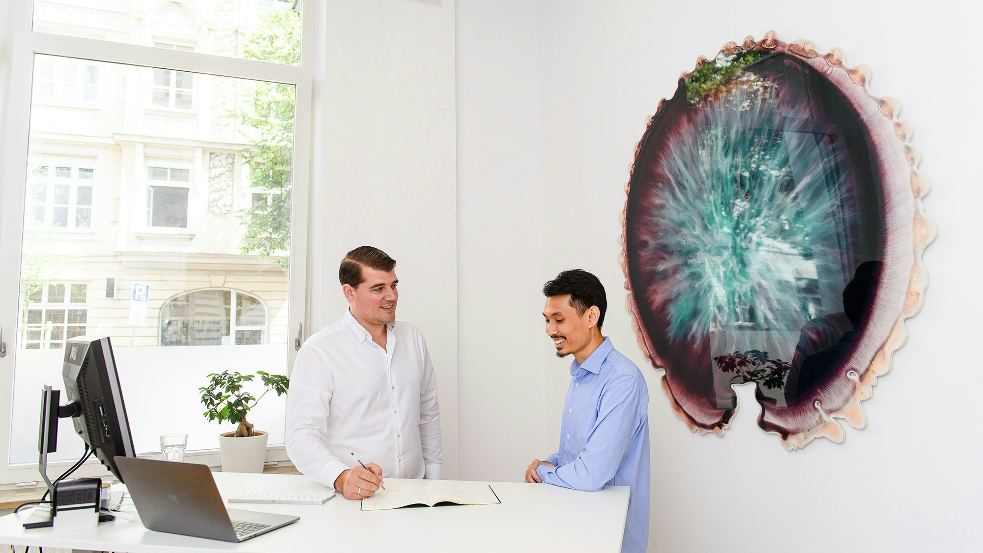 Zwei Kollegen unterhalten sich am Schreibtisch und im Hintergrund schmückt ein großes Kunstwerk in dunklen Farbtönen die Wand.