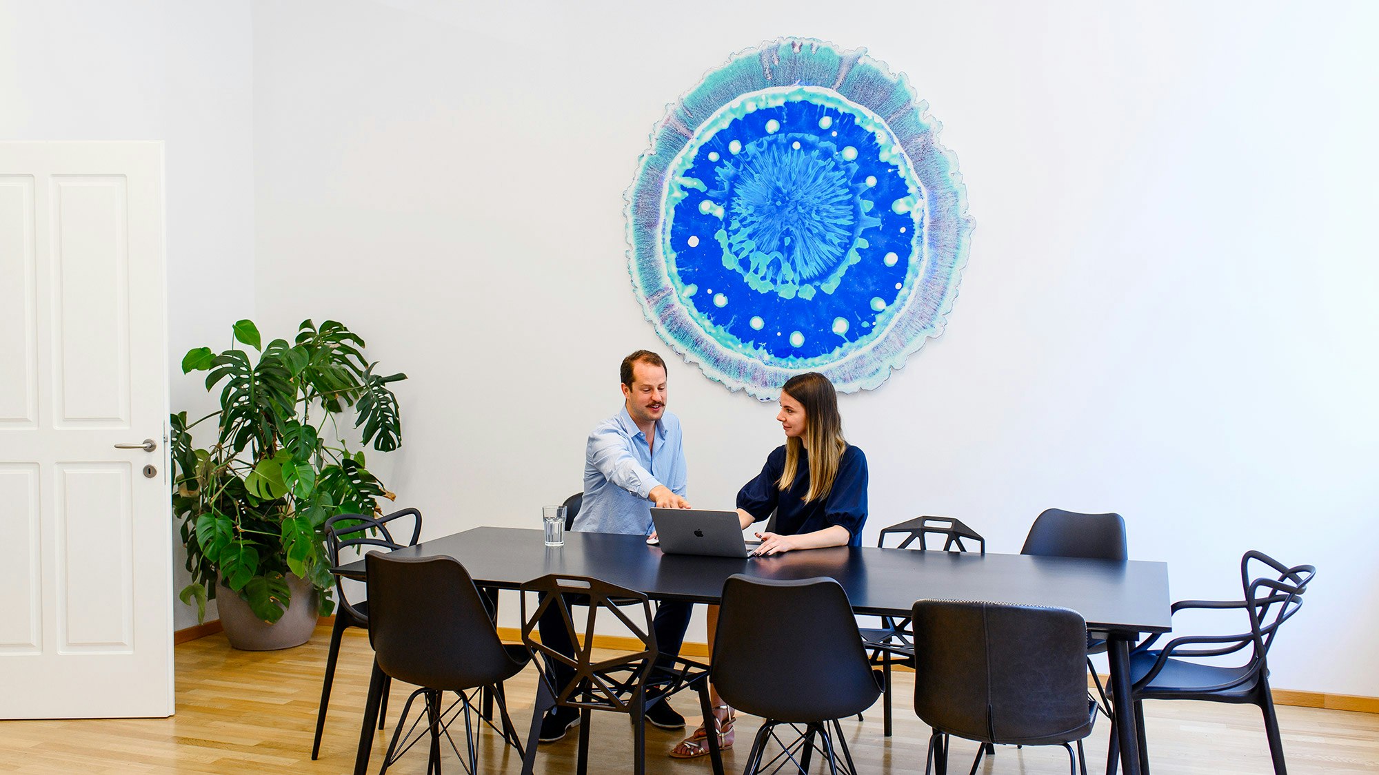 Two colleagues are sitting at the black meeting table and have a call on the laptop. In the background a large work of art in light blue.