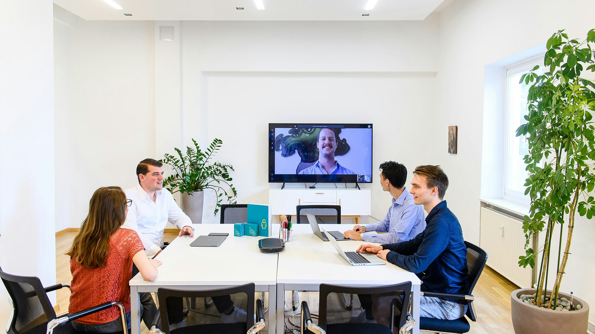 Ein Team aus vier Kollegen sitzt im Konferenzraum zusammen und führen einen Videocall der auf einem großen Bildschirm übertragen wird.