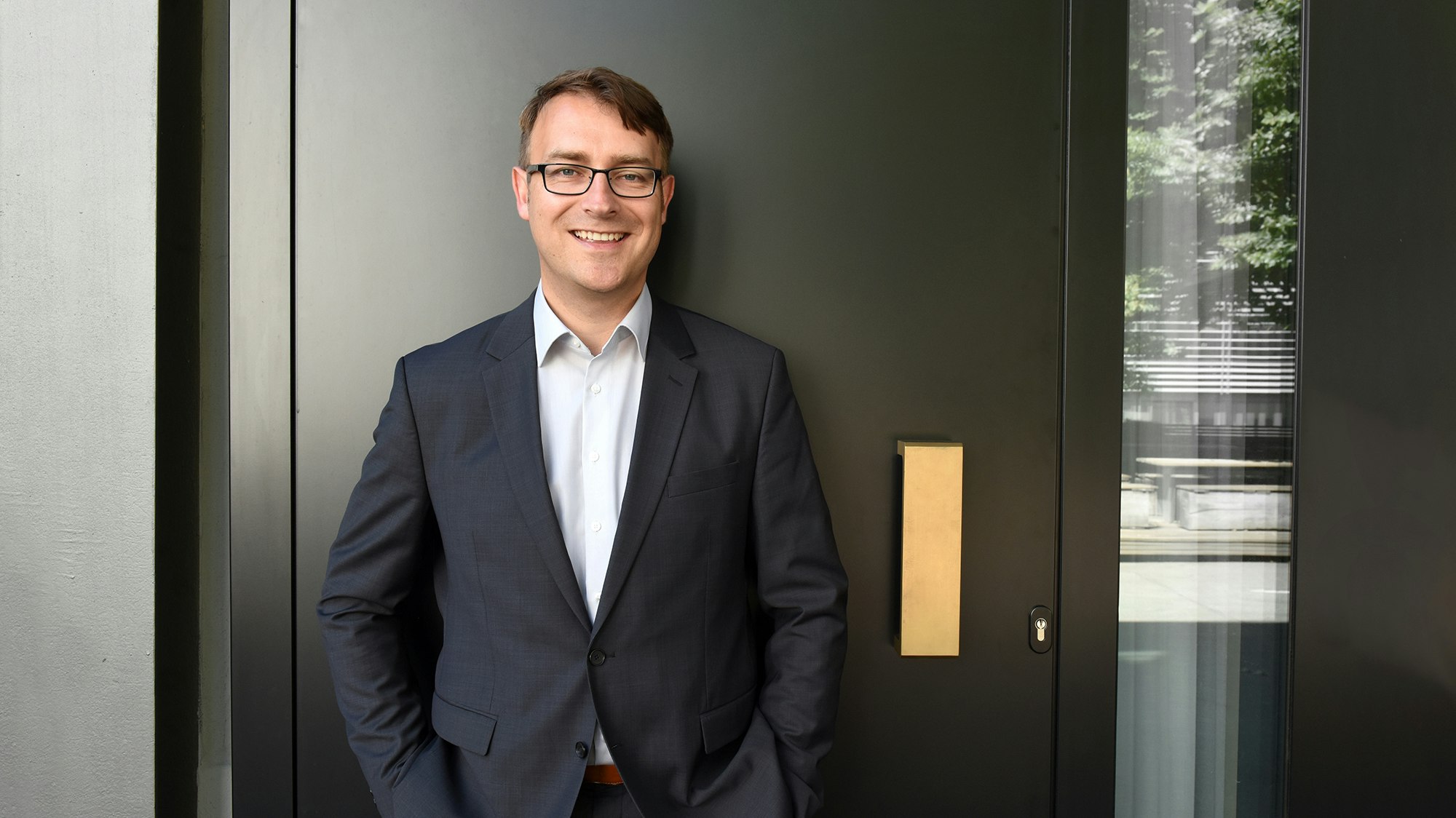 Kevin in a suit, leaning against a door and smiling