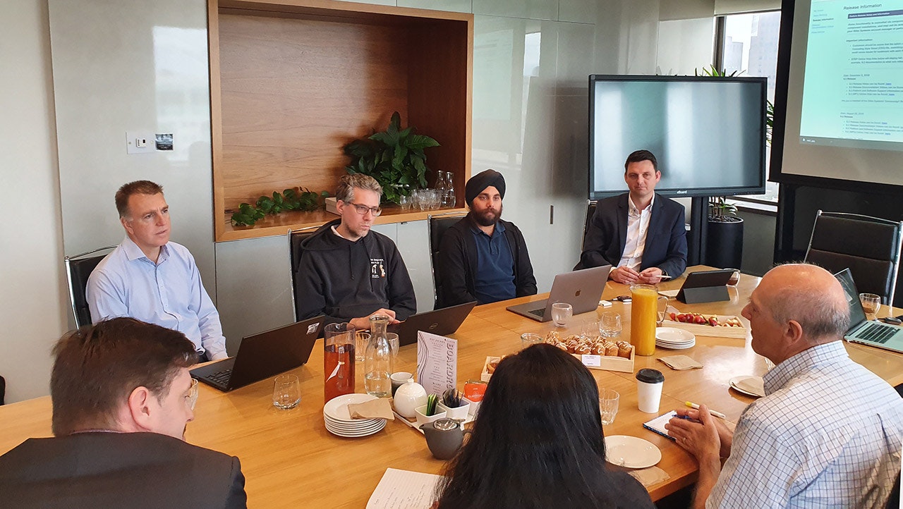 The team sits around a long table and discusses the topic. The Melbourne skyline can be seen in the background