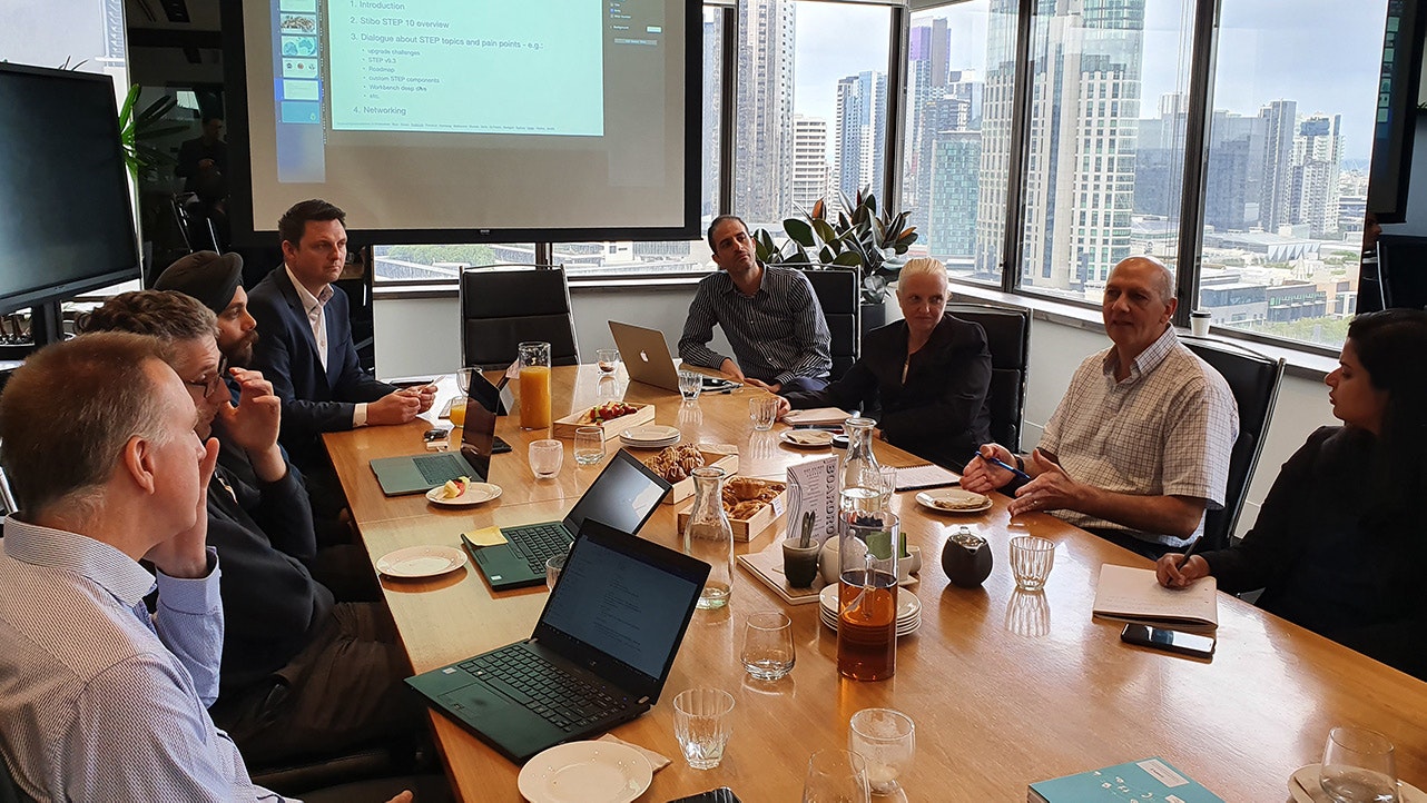The team sits around a long table and discusses the topic. The Melbourne skyline can be seen in the background