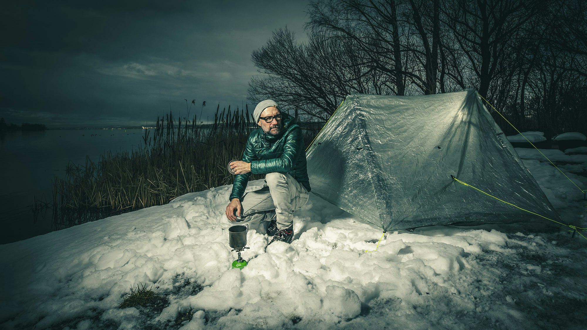 Man with jacket sits in the snow in front of the tent and makes tea on the gas stove.