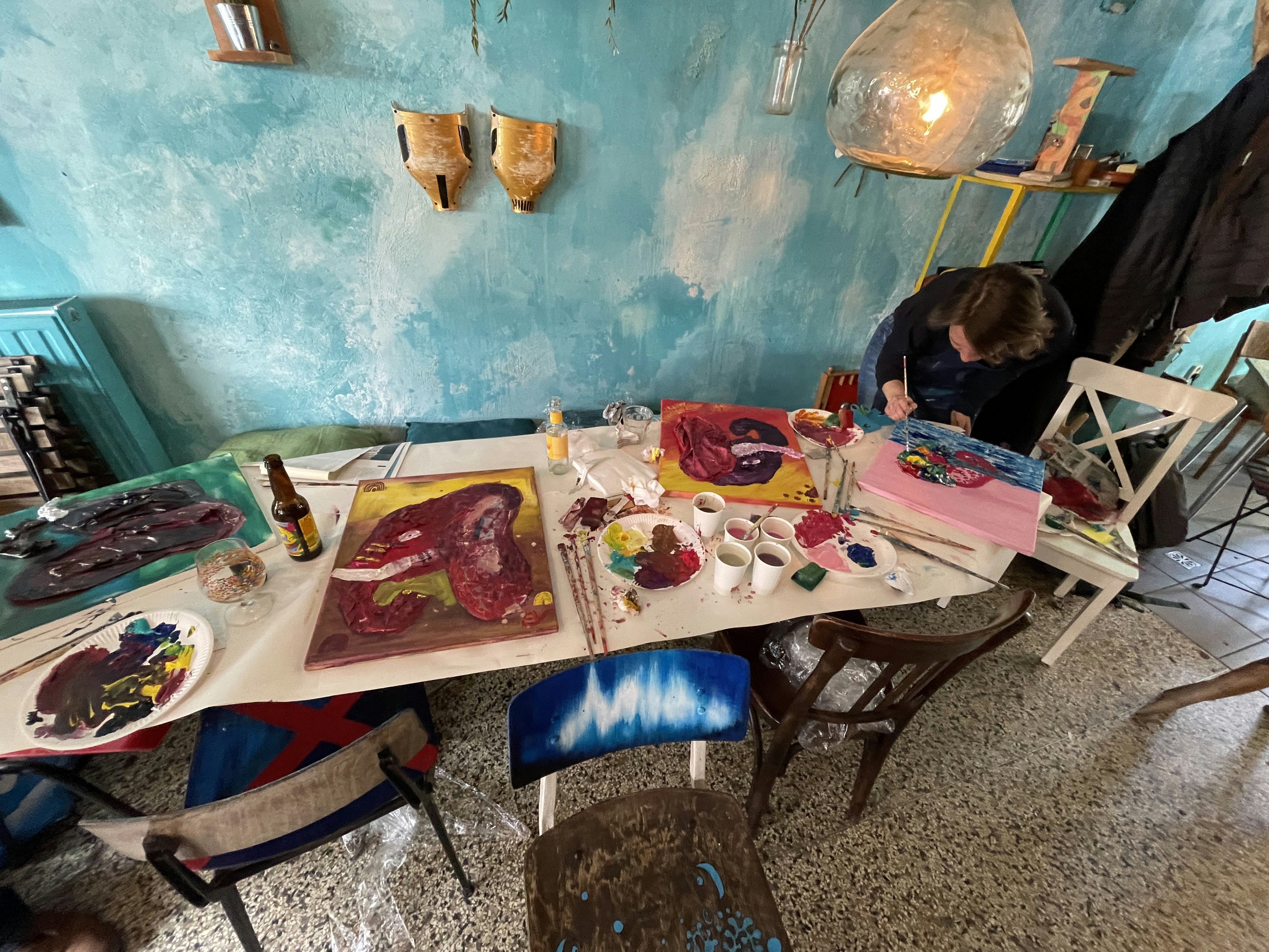 Finished mixed media paintings of elephants on a table together with office supplies and a person finalizing their canvas.