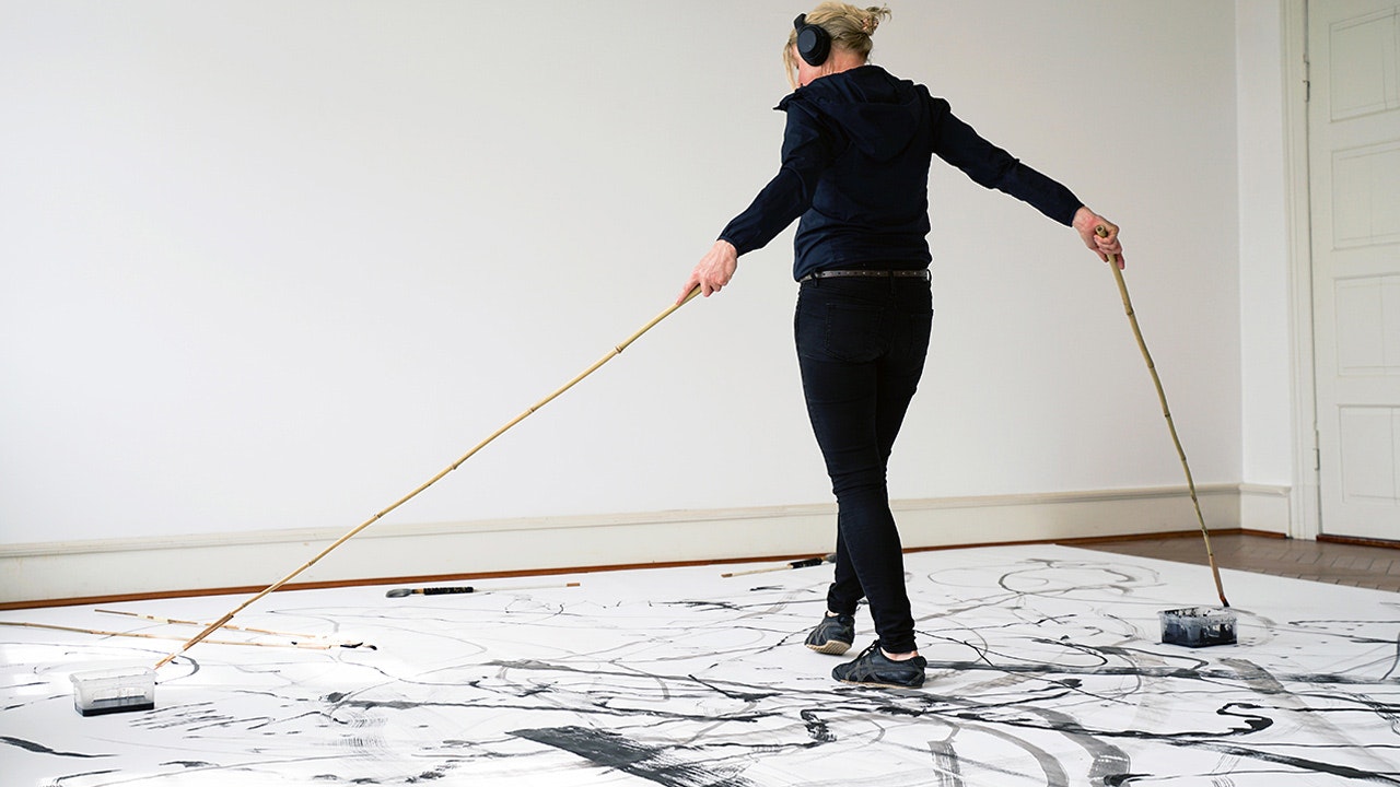Künstlerin tanzt auf ihrer am Boden liegenden Leinwand mit zwei langen Pinseln und zeichnet Formen, während sie mit Kopfhörern Musik hört.