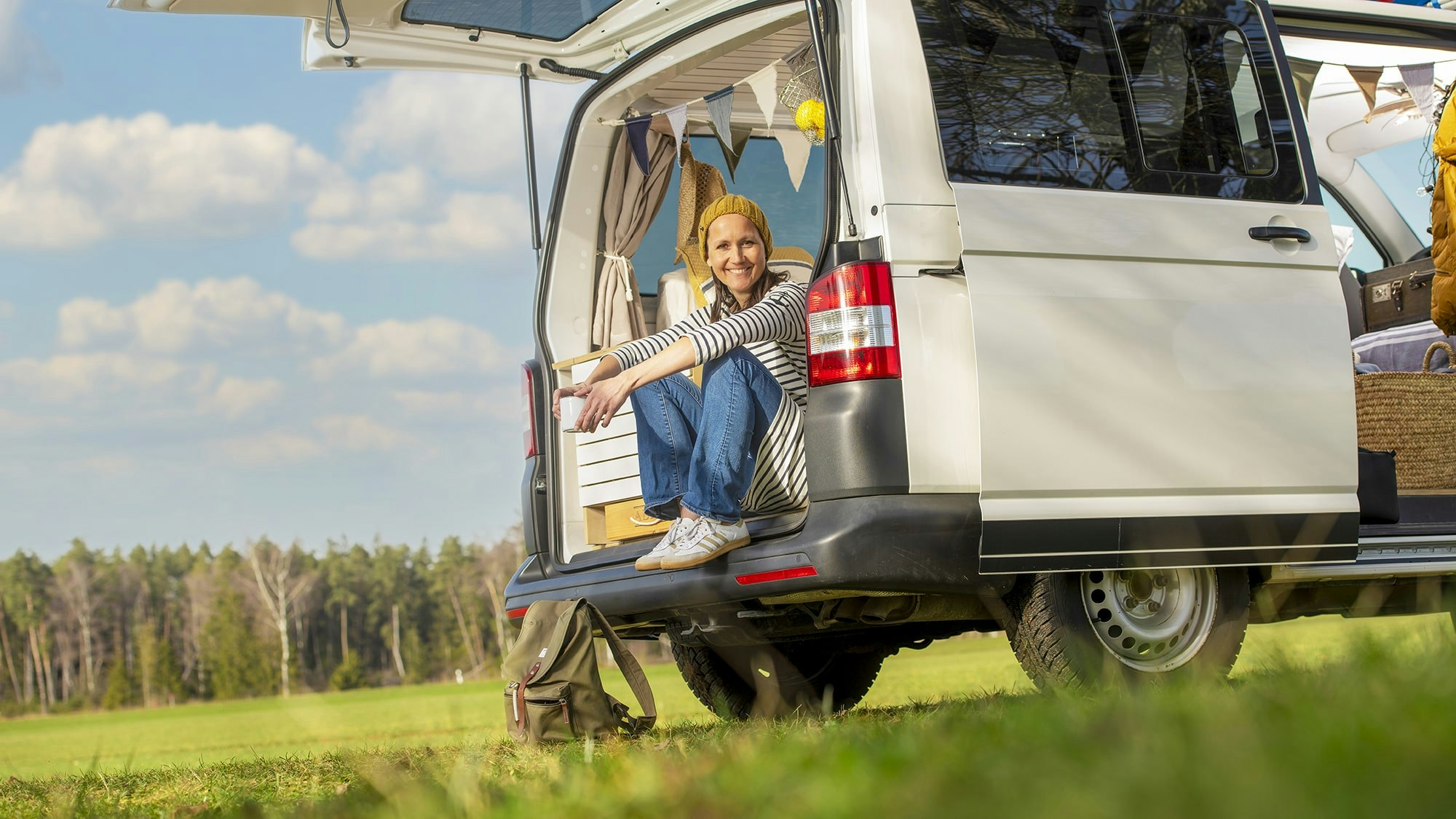 Woman with wool cap sitting outside in nature in camping bus with open back door.