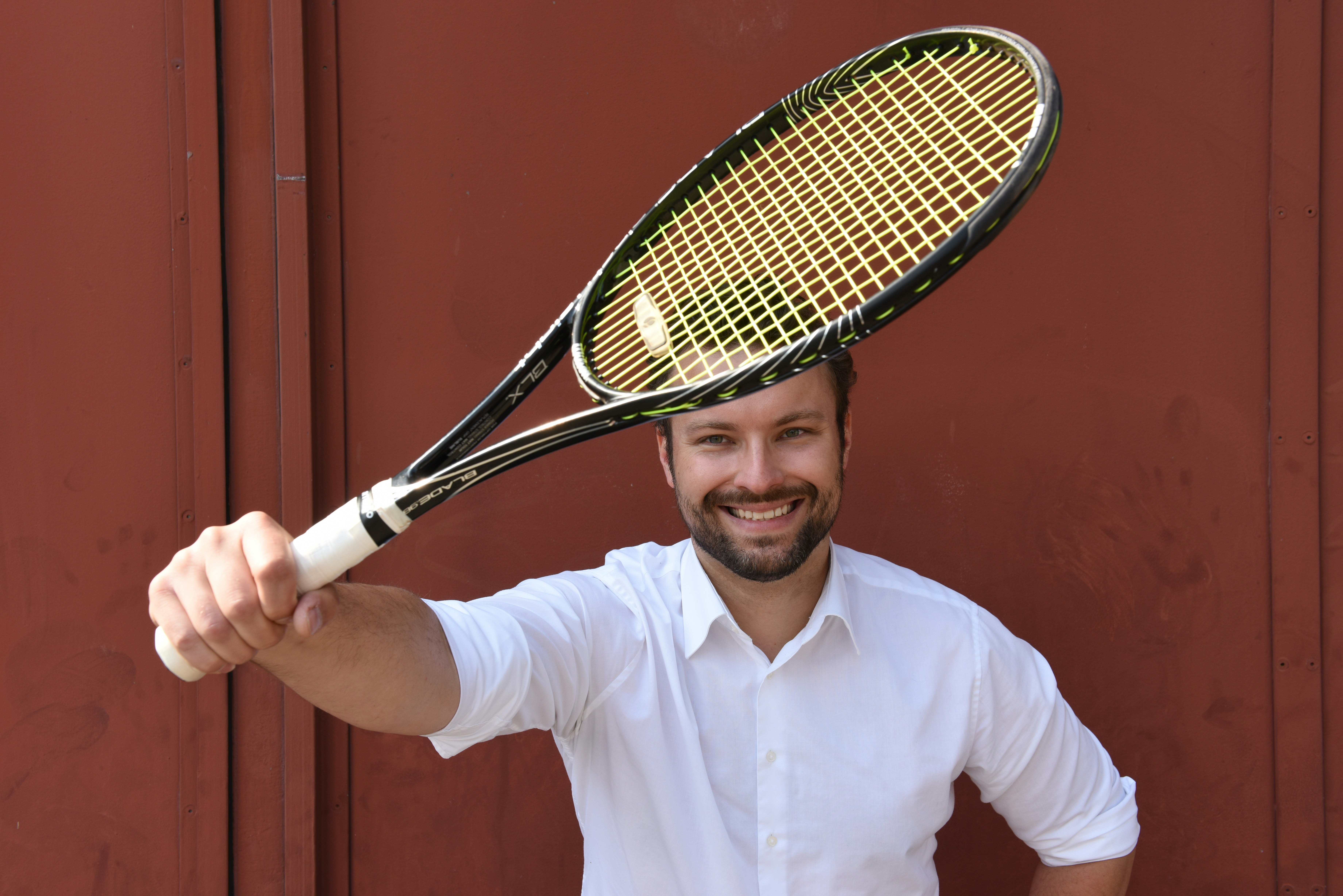 Person smiling and holding a tennis racket infront of a red wall