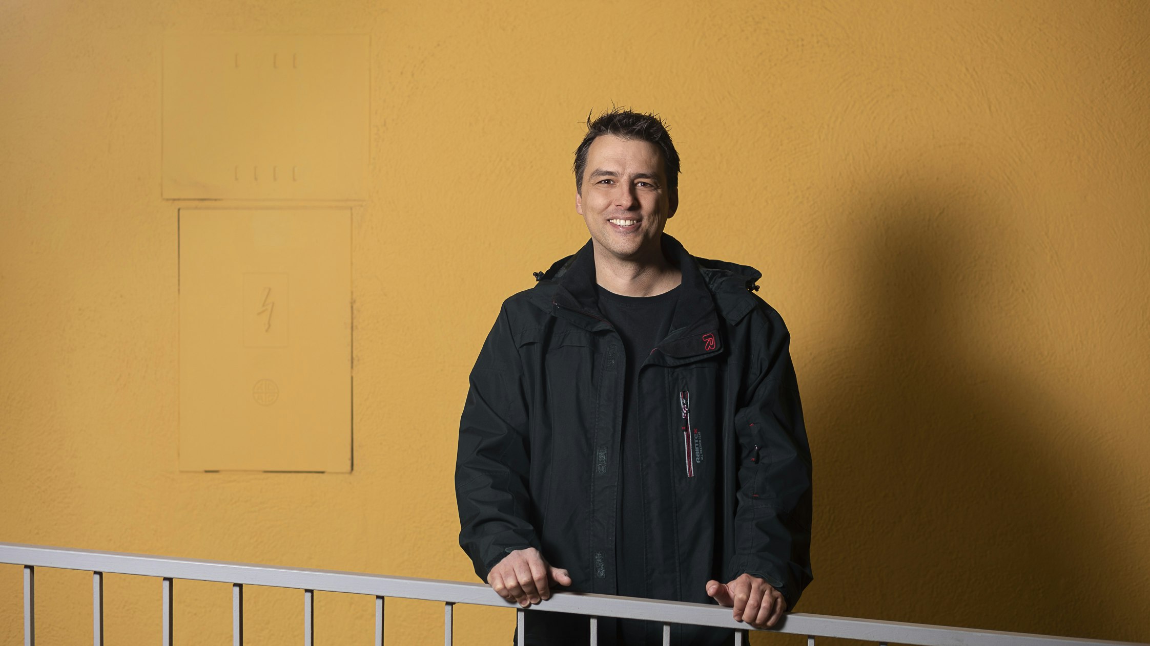 Georg laughs into the camera, the background is in a rich mustard yellow.