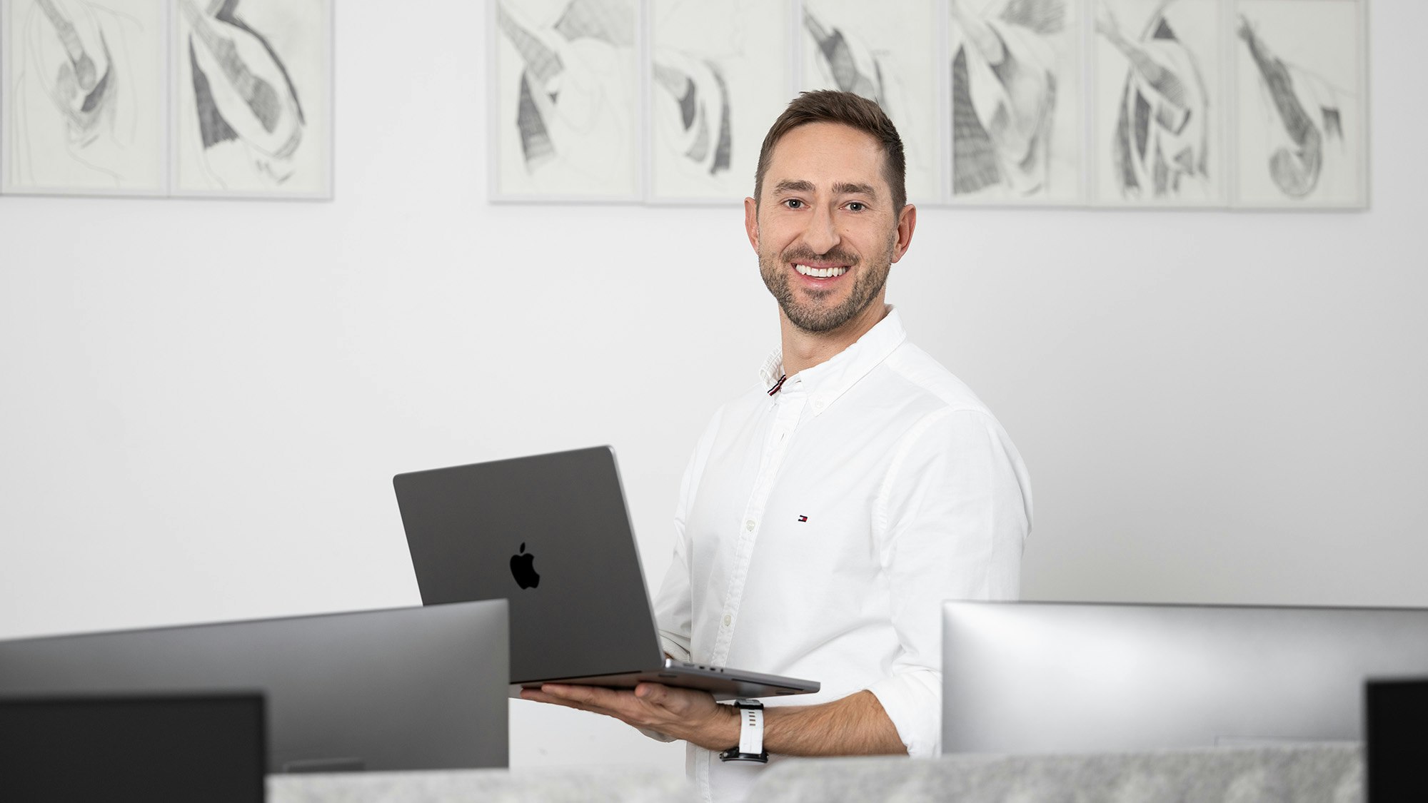 Thomas steht mit seinem Laptop vor einer Kunstwerkreihe.